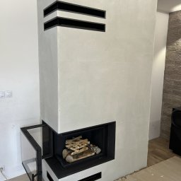 HYDRO-TECH RAFAŁ JAWORSKI - Nowoczesny kominek z otwartymi drzwiczkami, wewnątrz ułożone drewno. Biała obudowa z czarnymi, poziomymi akcentami. Widoczne niedokończone wykończenie podłogi.