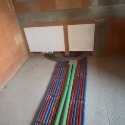 HYDRO-TECH RAFAŁ JAWORSKI - Instalacja ogrzewania podłogowego w trakcie montażu: rury grzewcze w kolorach czerwonym i niebieskim ułożone równolegle, zielone rury wentylacyjne, surowa cegła.