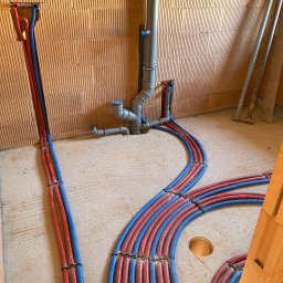 HYDRO-TECH RAFAŁ JAWORSKI - Instalacja ogrzewania podłogowego w trakcie montażu: czerwono-niebieskie rury ułożone na podłożu, widoczne elementy mocujące i pionowe rury kanalizacyjne przy ścianie z cegły.