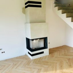 HYDRO-TECH RAFAŁ JAWORSKI - Nowoczesny kominek narożny z czarnymi akcentami, jasne drewno na podłodze ułożone w jodełkę, surowe schody betonowe w tle, minimalistyczny design.