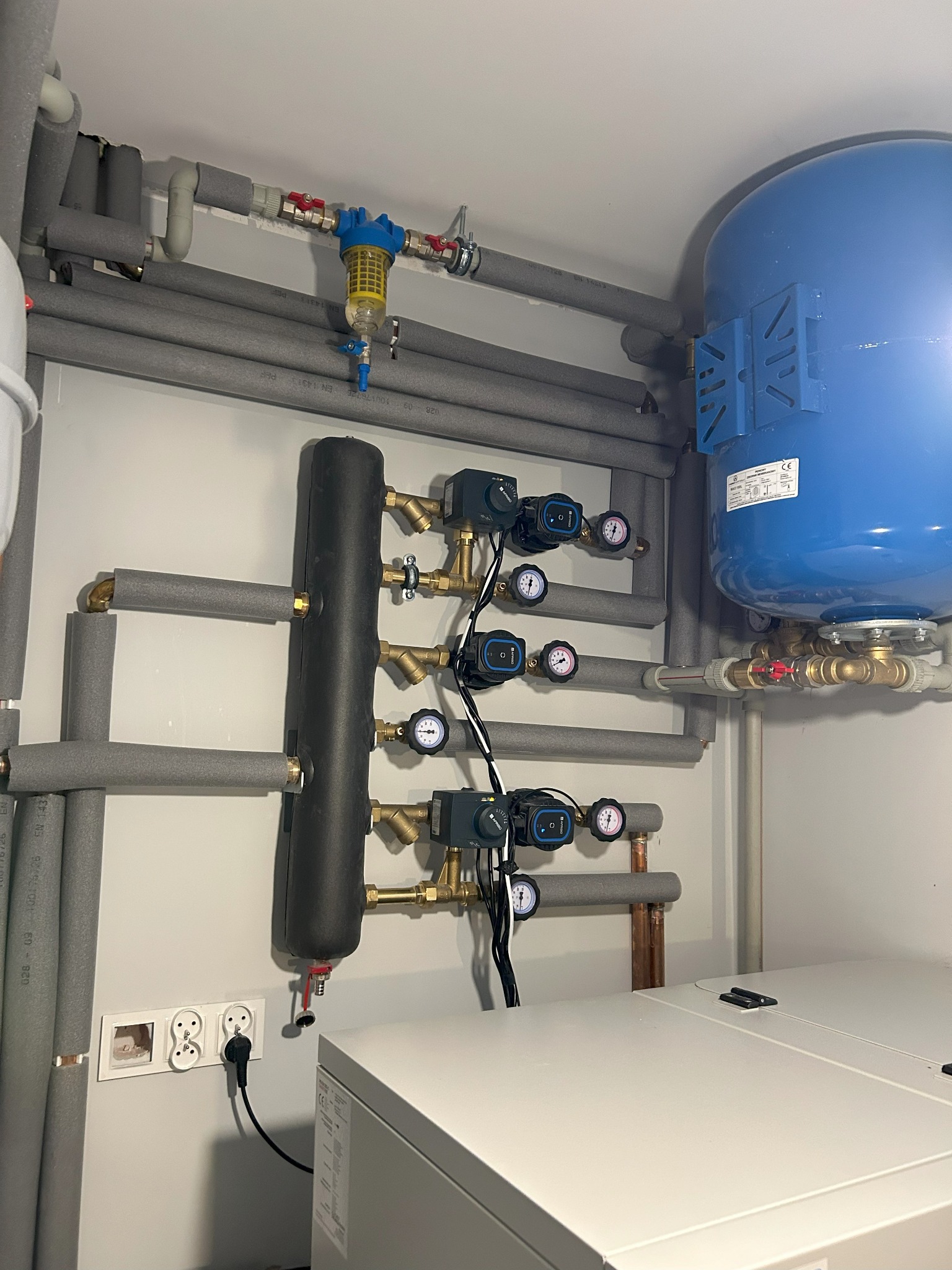 Instalacja grzewcza w kotłowni: niebieski zbiornik, czarny filtr, liczne rury z izolacją termiczną, manometry i pompy obiegowe na białej ścianie, widoczne gniazdka elektryczne i fragment kotła.