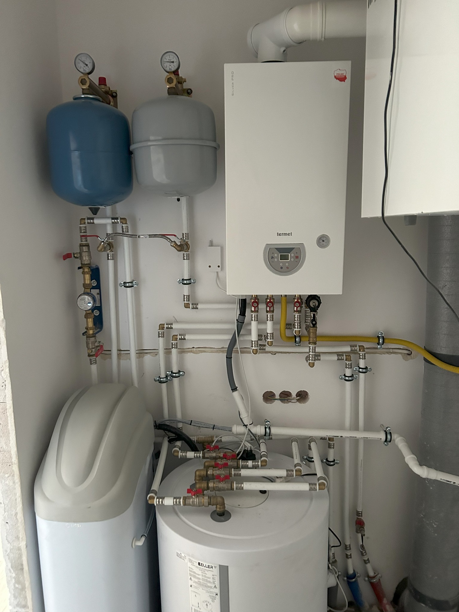 Biały piec gazowy Termet z podłączonymi rurami gazowymi i wodnymi, obok zbiornik hydroforowy w kolorze niebieskim i szarym, oraz biały zmiękczacz wody Keller, całość zamontowana na białej ścianie.