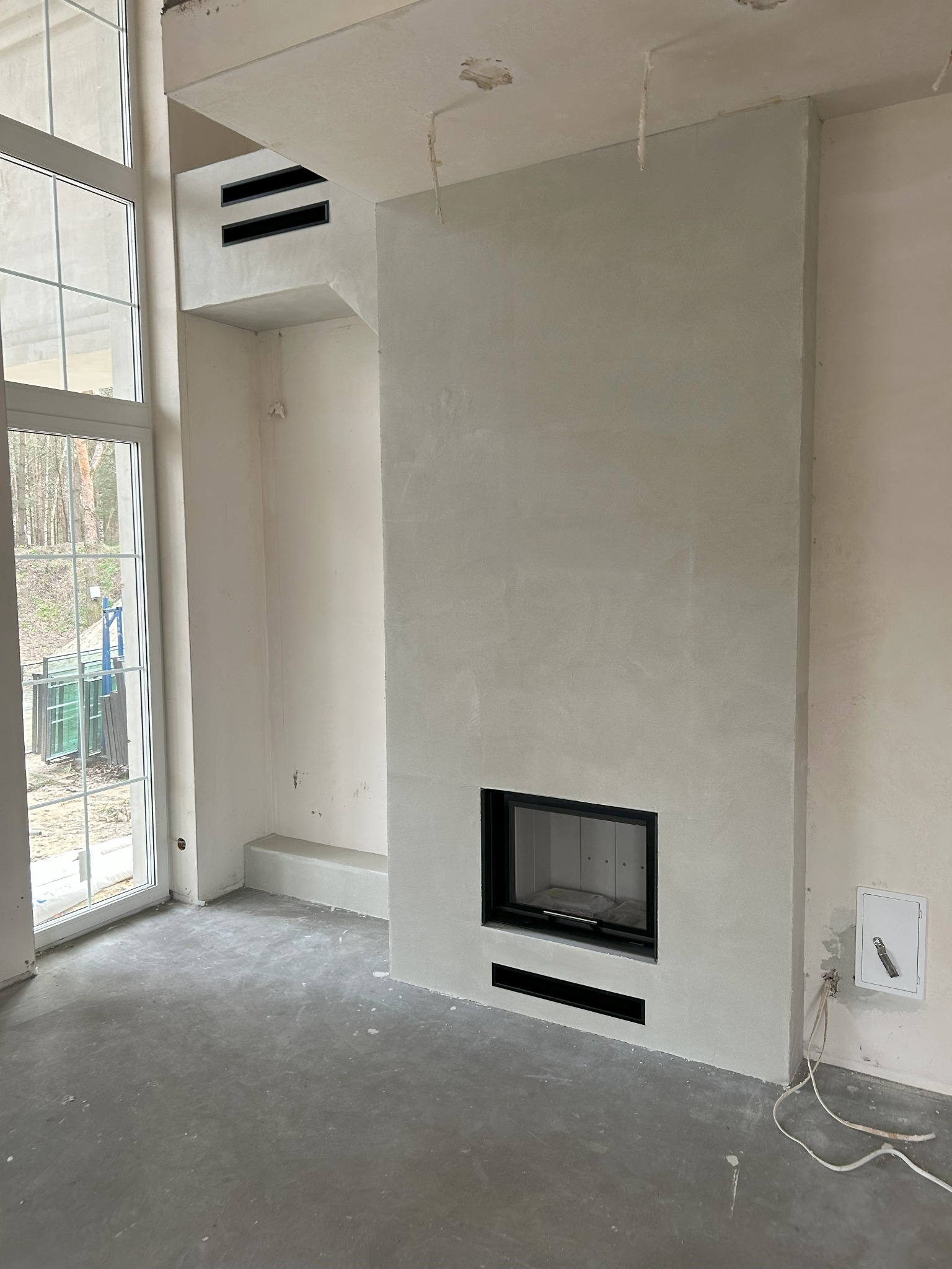 Surowy stan wykończenia wnętrza z wnęką kominkową, widoczne otwory wentylacyjne nad kominkiem, okno z widokiem na drzewa, instalacja elektryczna wystająca ze ściany.