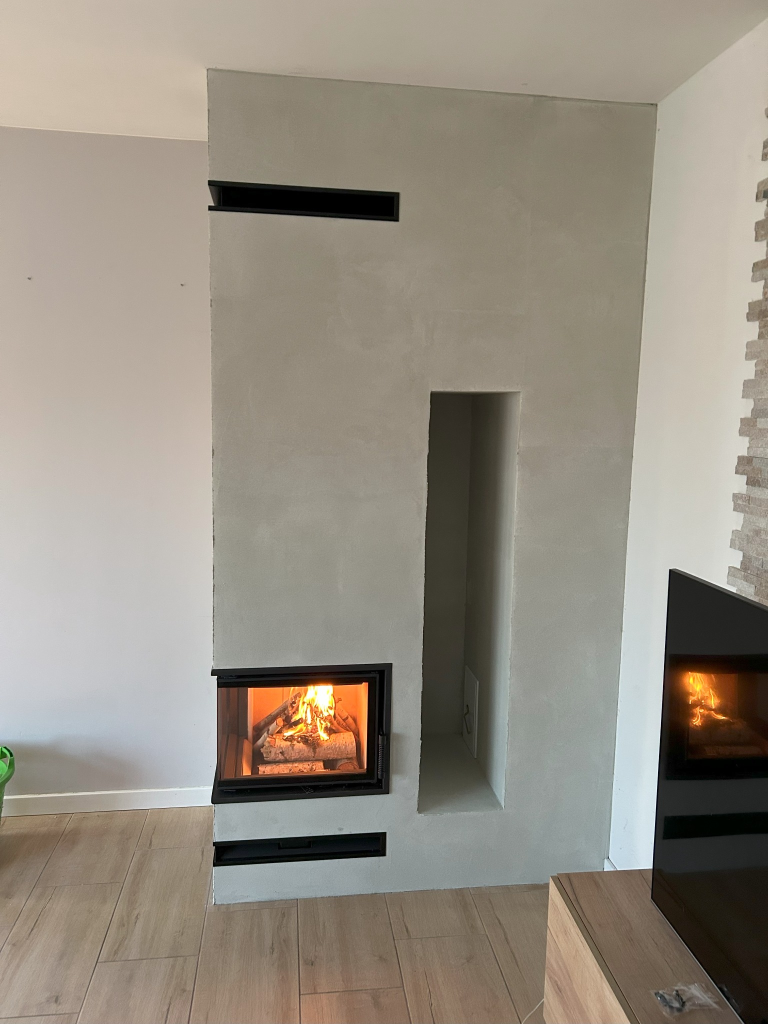 Nowoczesny kominek z szarą obudową, płomień widoczny przez czarną ramę, drewniana podłoga i fragment ściany z dekoracyjnym kamieniem w tle.