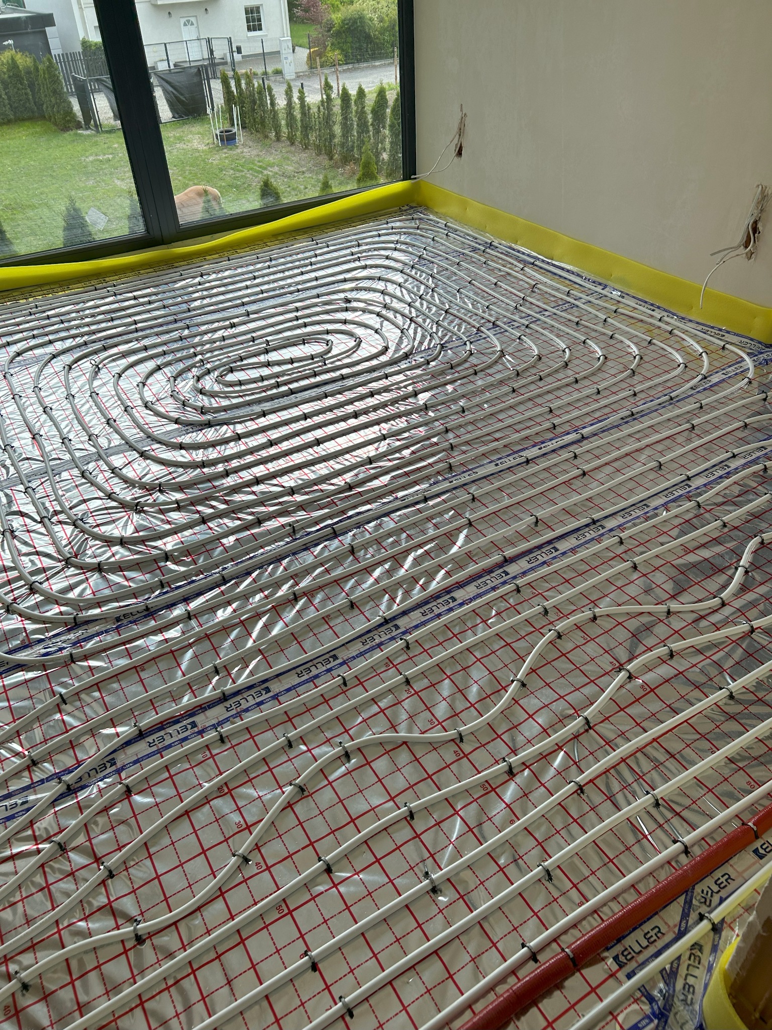 Instalacja ogrzewania podłogowego z rur Keller na folii z siatką, widok z góry. Żółta taśma brzegowa, okno z widokiem na ogród. Etap przed wylewką.