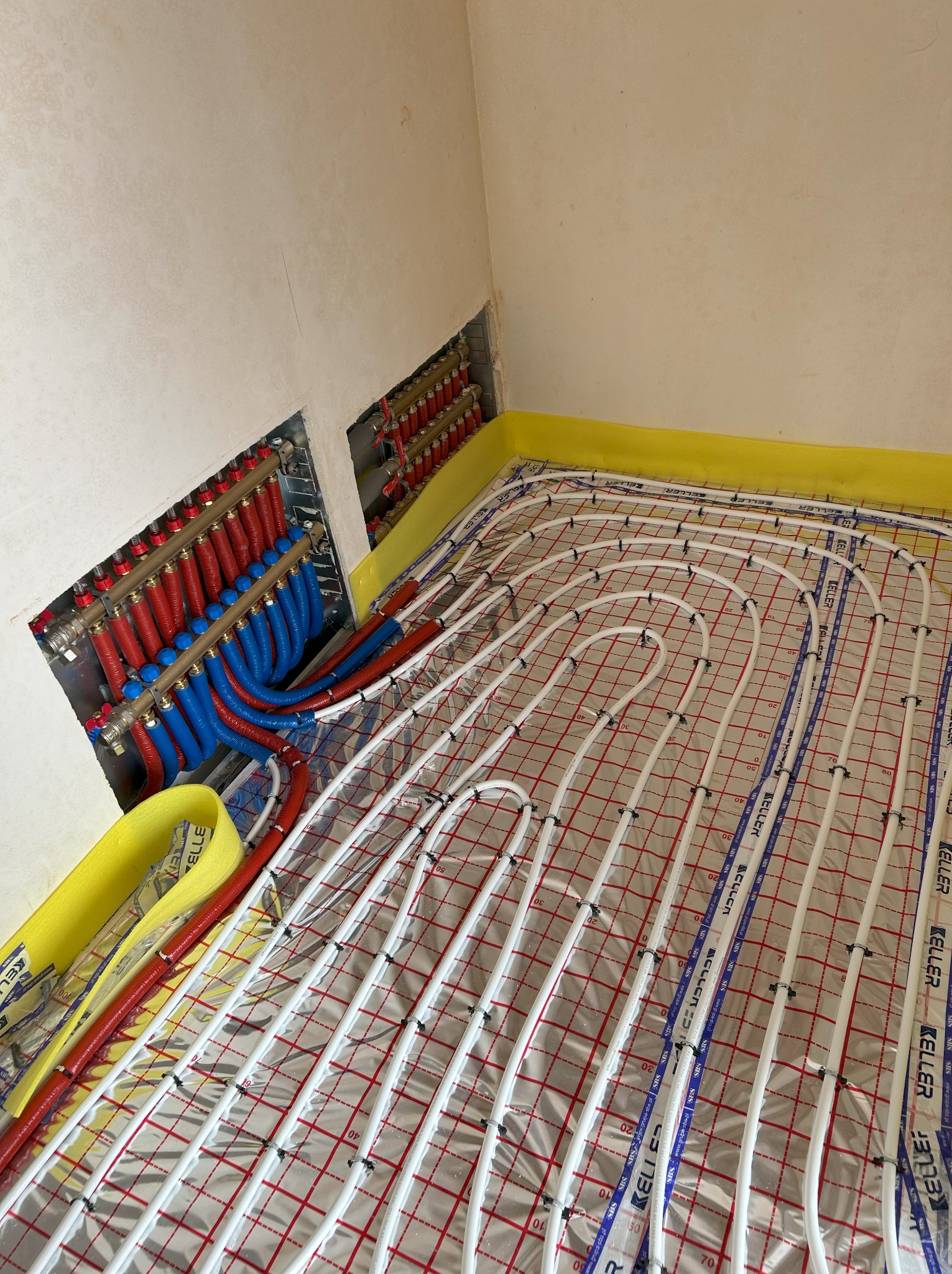 Instalacja ogrzewania podłogowego z rozdzielaczem w ścianie. Białe rurki układane na folii z czerwoną siatką i niebieskimi oznaczeniami.