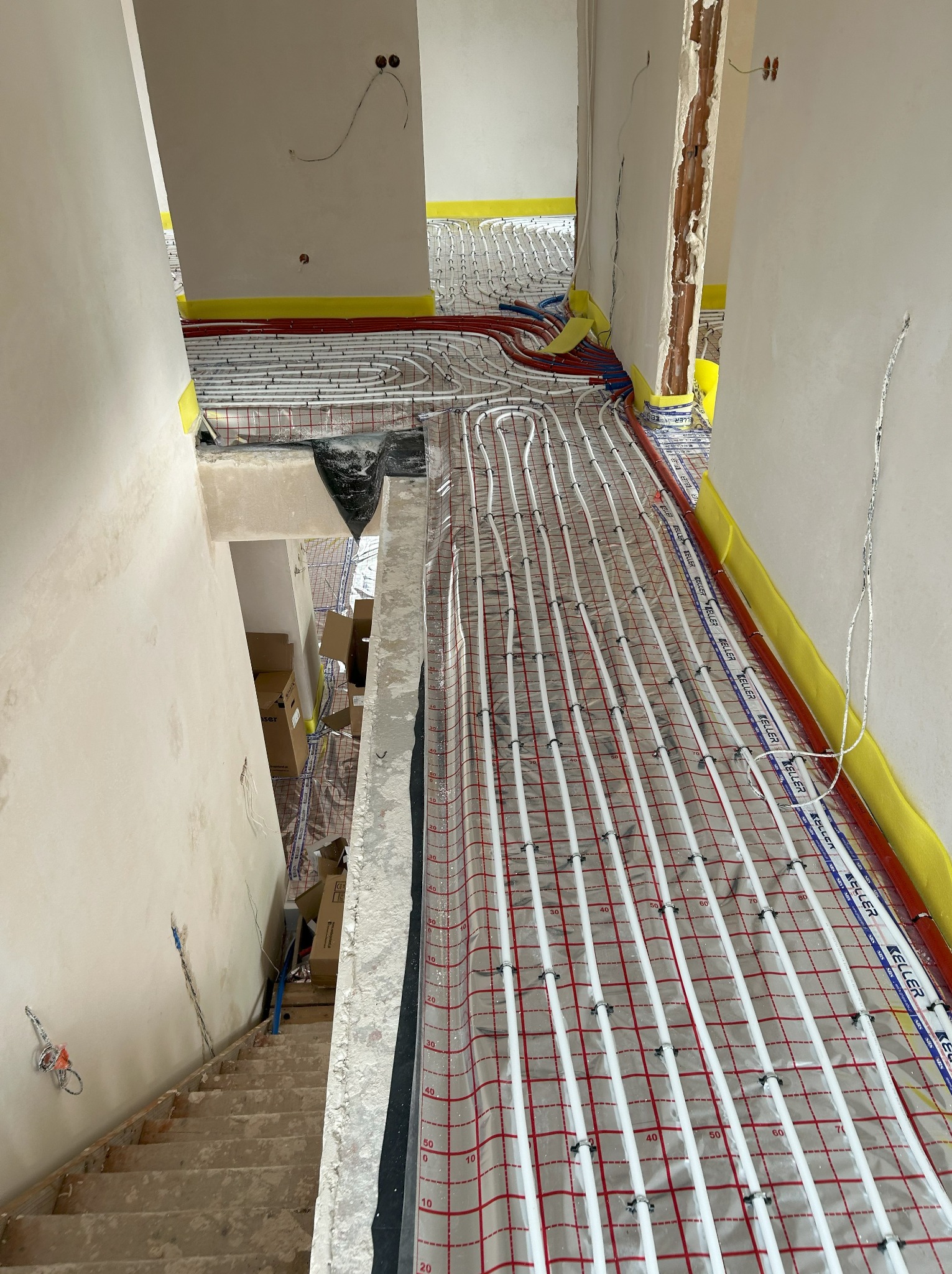 Instalacja ogrzewania podłogowego z białych rur na folii z siatką i oznaczeniami co 10 cm, zabezpieczona żółtą taśmą przy ścianach w surowym wnętrzu.