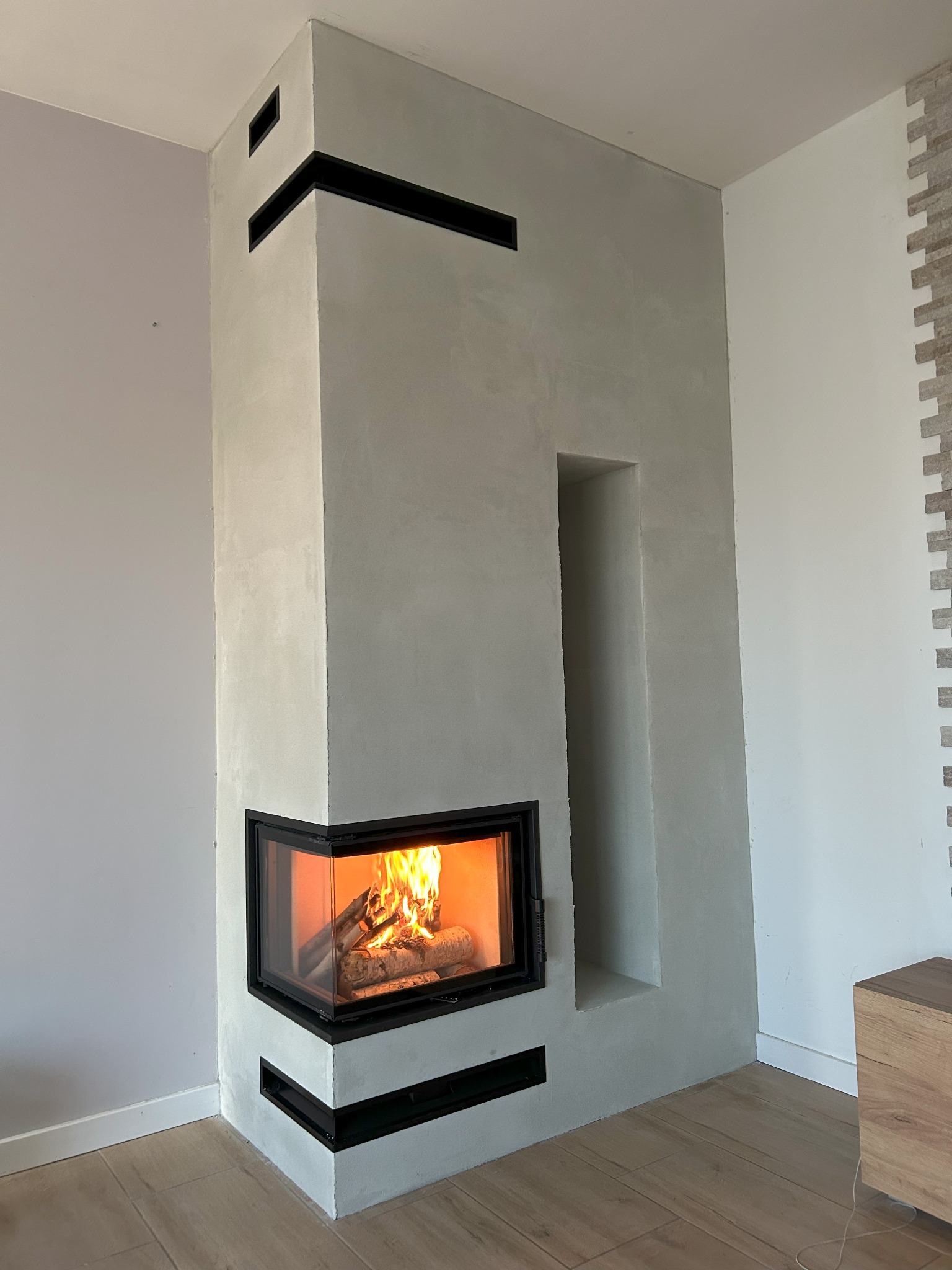 Nowoczesny kominek narożny z paleniskiem, obudowany szarym tynkiem z czarnymi akcentami. Widoczne płomienie i drewno wewnątrz. Minimalistyczny design.