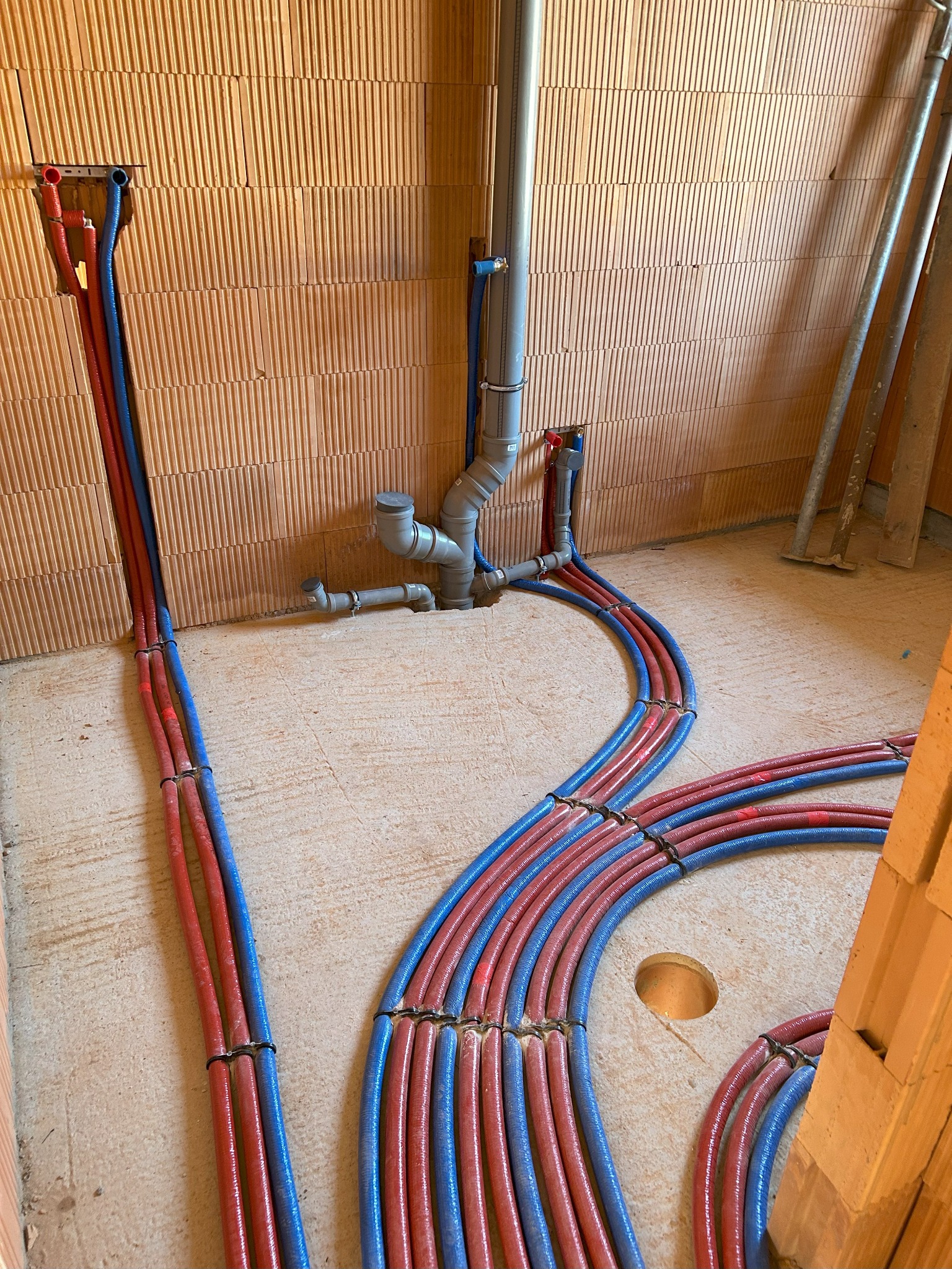 Instalacja wodna podłogowa w trakcie montażu: rury grzewcze ułożone w pętle na podłożu, widoczne przyłącza do ściany z pustaków ceramicznych.