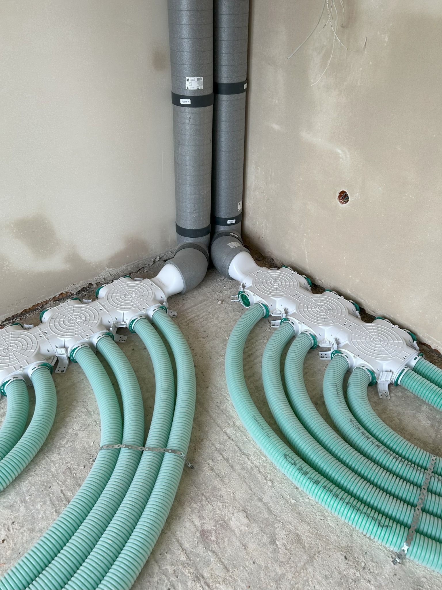 Szare rury wentylacyjne i zielone, elastyczne przewody w rozdzielaczu na surowym betonie, w rogu pomieszczenia. Instalacja rekuperacji w trakcie montażu.