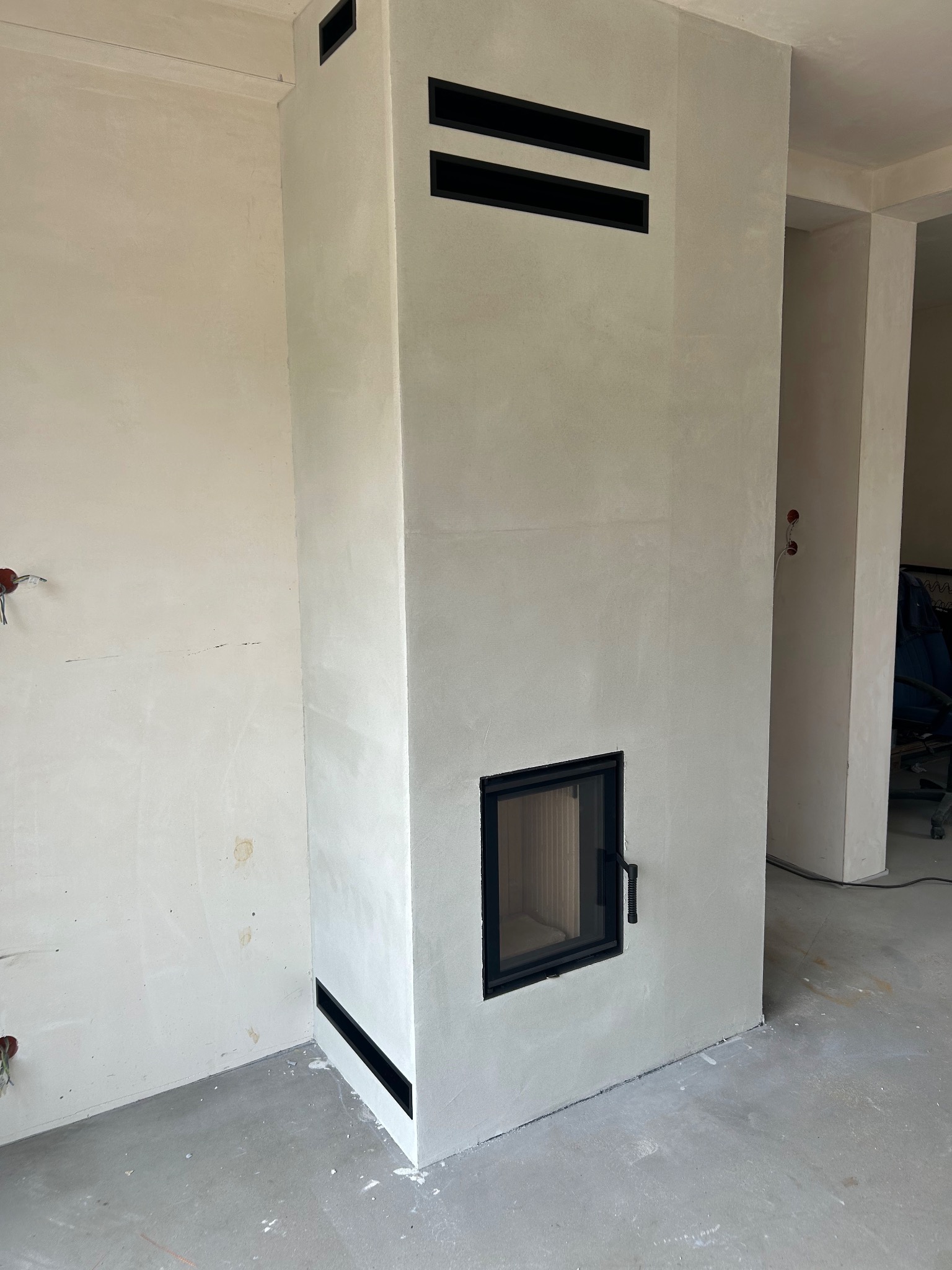 Nowoczesny, kremowy kominek z czarnymi, poziomymi wlotami powietrza i szybą, w surowym wnętrzu. Widoczne niedoskonałości ścian, co sugeruje etap wykończenia.