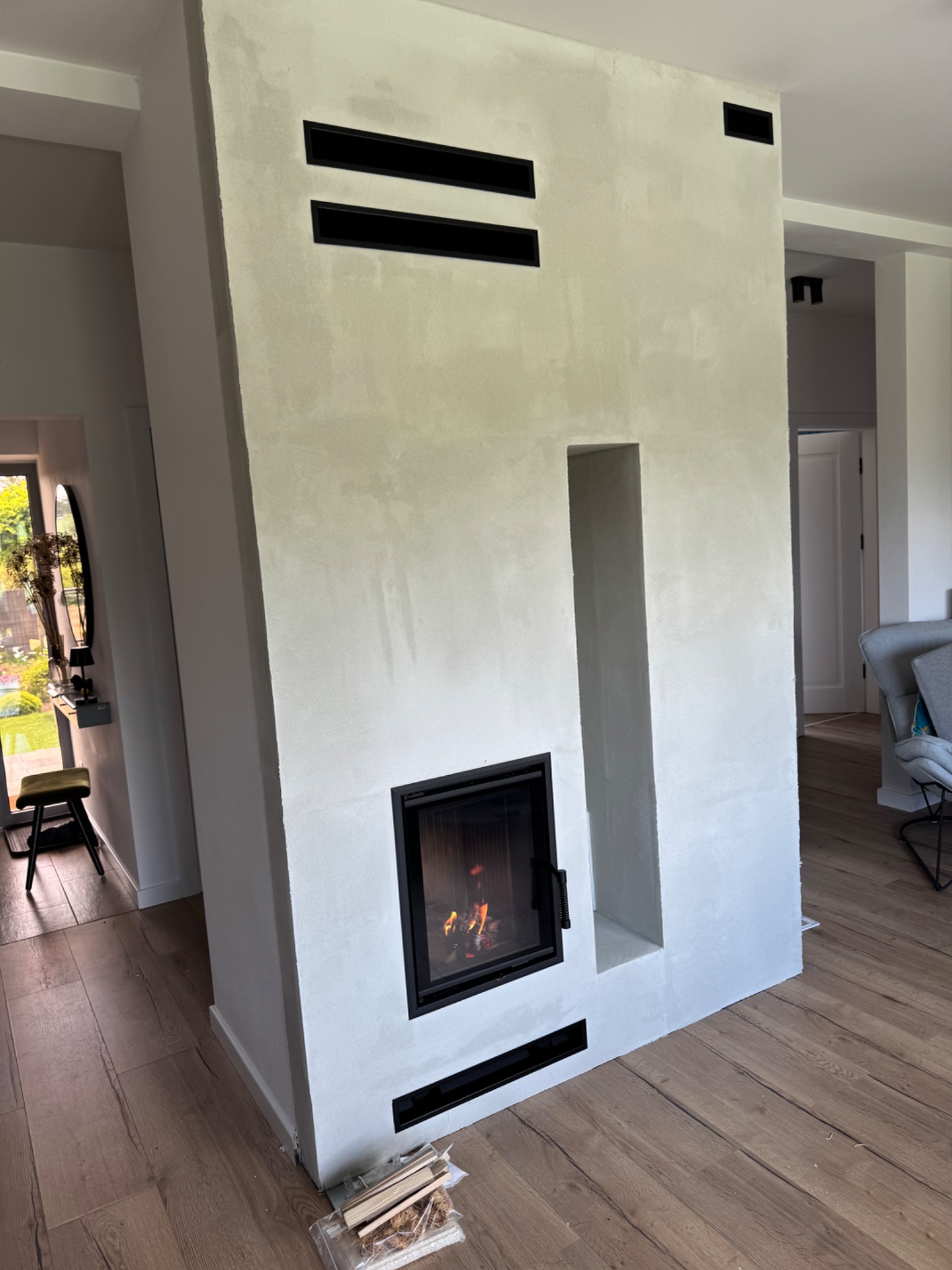 Nowoczesny kominek z paleniskiem w obudowie z szarego betonu architektonicznego, z czarnymi wlotami powietrza. Ogień w palenisku. Parkiet na podłodze.