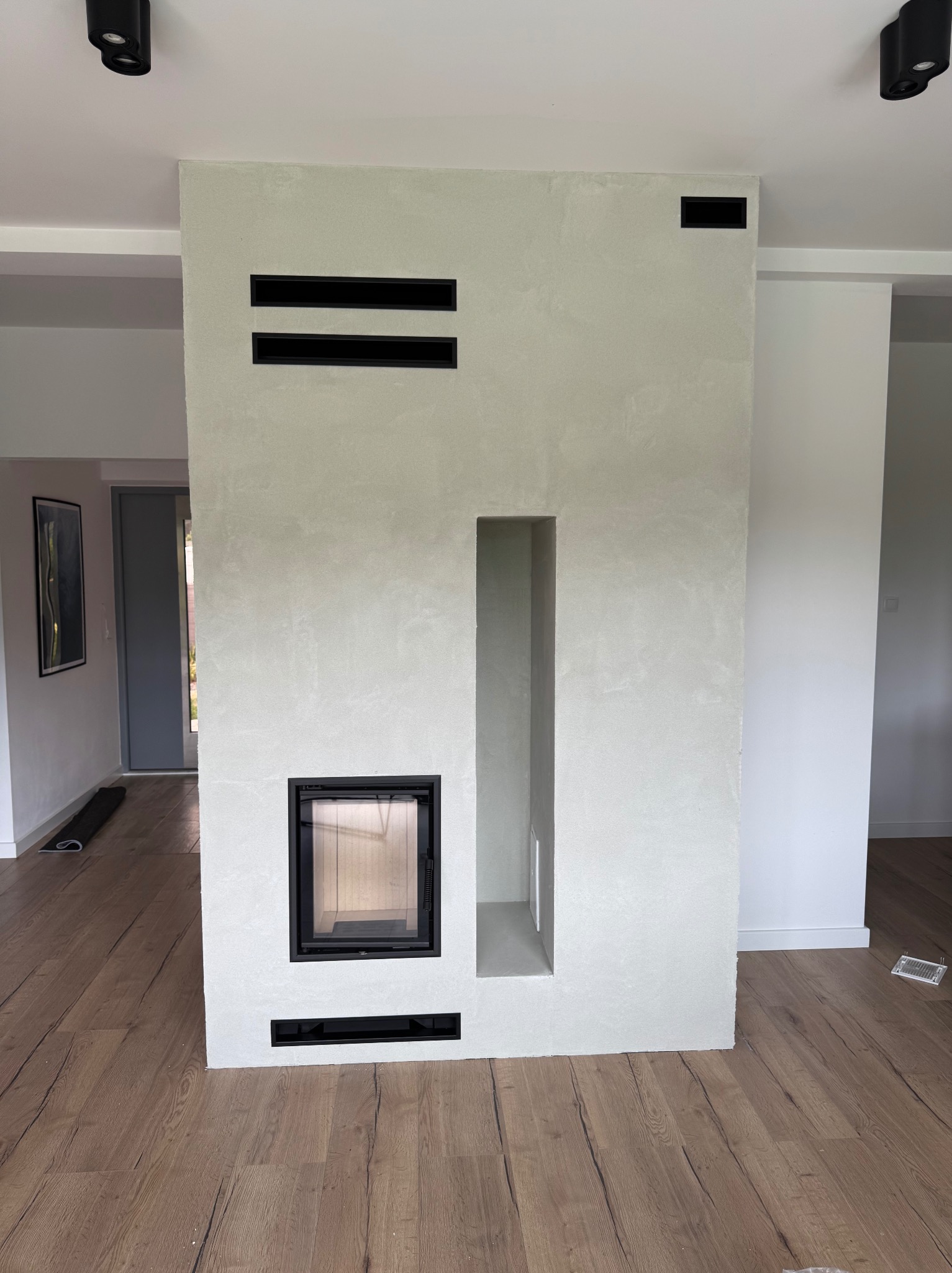Nowoczesna obudowa kominkowa w jasnym odcieniu, z czarnymi elementami wentylacyjnymi, w minimalistycznym wnętrzu z drewnianą podłogą i oświetleniem punktowym.