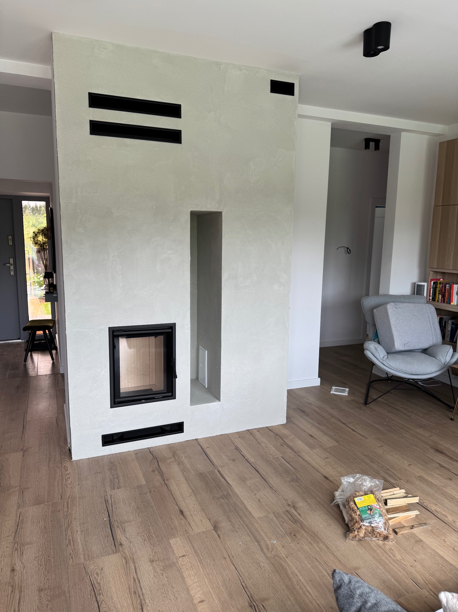 Nowoczesny kominek z czarnymi akcentami w Nadarzynie. Minimalistyczny design, jasne wykończenie ściany, drewniana podłoga. Widoczne drewno do kominka.