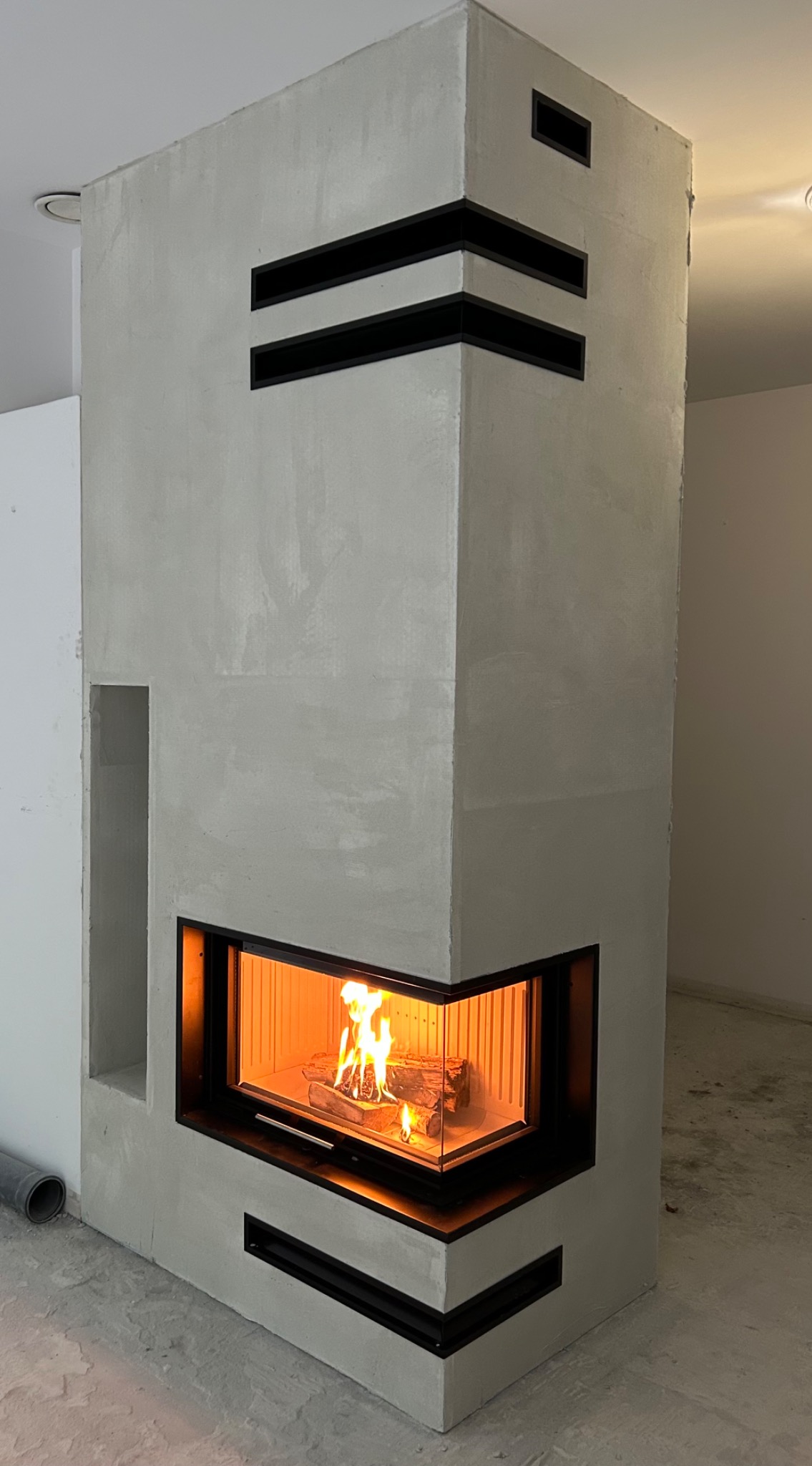 Nowoczesny kominek narożny z płomieniami, obudowany szarym tynkiem strukturalnym i czarnymi detalami, w surowym wnętrzu.