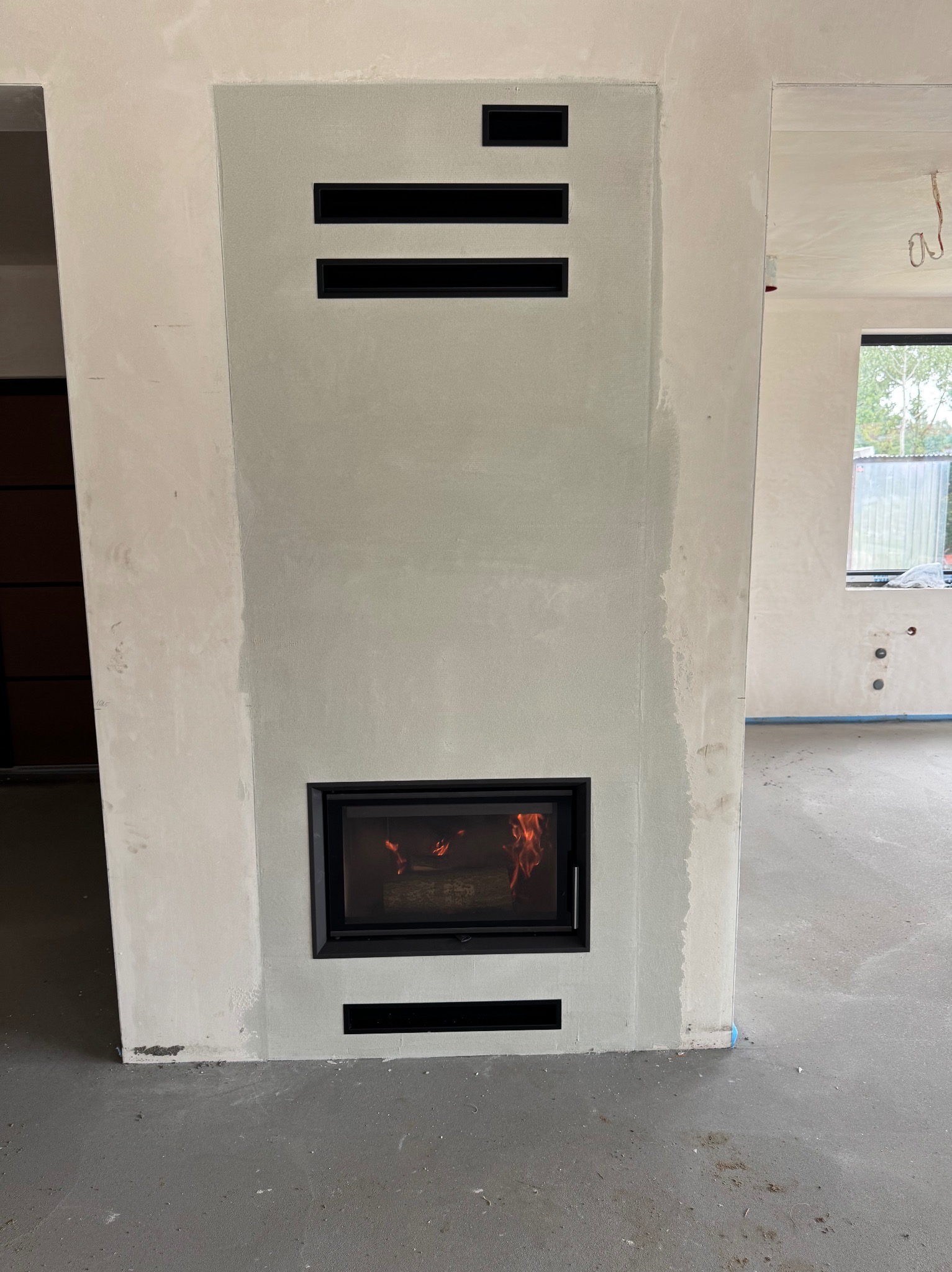 Wnętrze w trakcie wykończenia z wbudowanym kominkiem z czarną obudową i palącym się ogniem. Widoczne otwory wentylacyjne w obudowie kominka.