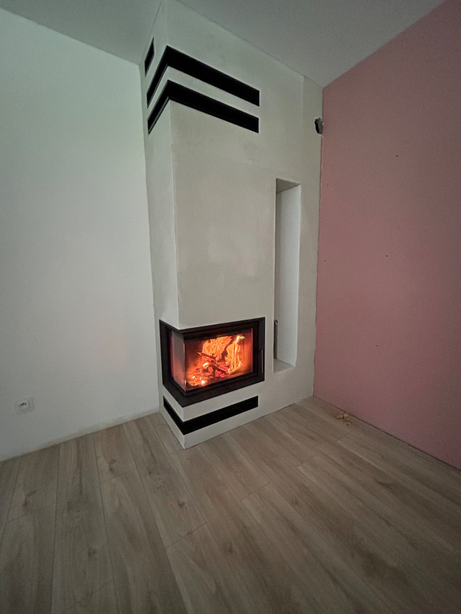 Nowoczesny kominek narożny z paleniskiem, obudowany białym kamieniem z czarnymi akcentami, w jasnym wnętrzu z drewnianą podłogą i różową ścianą.