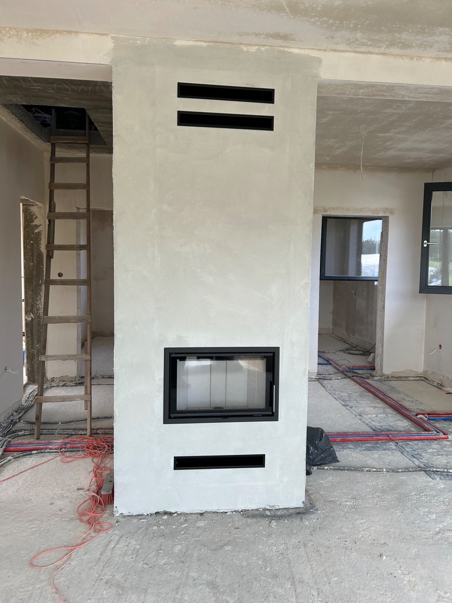 Nowoczesny kominek z czarnym wkładem, w surowym wnętrzu w trakcie wykończenia. Widoczne instalacje podłogowe i drabina. Minimalistyczny design kominka.