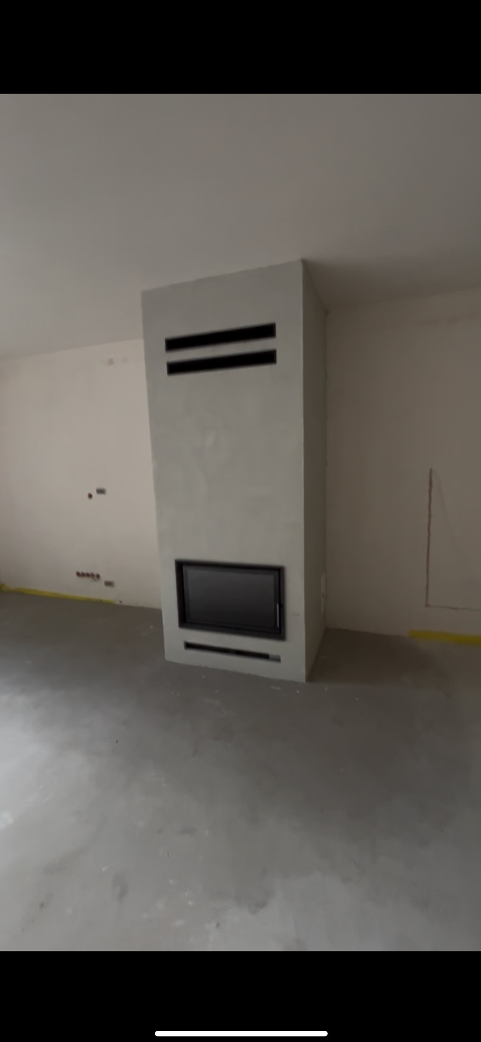 Nowoczesny kominek z czarnym wkładem w surowym, szarym wykończeniu w pustym pomieszczeniu. Minimalistyczny design z poziomymi szczelinami wentylacyjnymi.