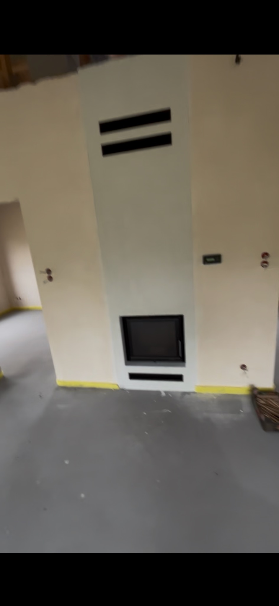 Nowoczesny kominek z czarnym wkładem, obudowany jasną płytą, z poziomymi szczelinami wentylacyjnymi. Żółta taśma malarska na podłodze. Widok na surowy pokój.