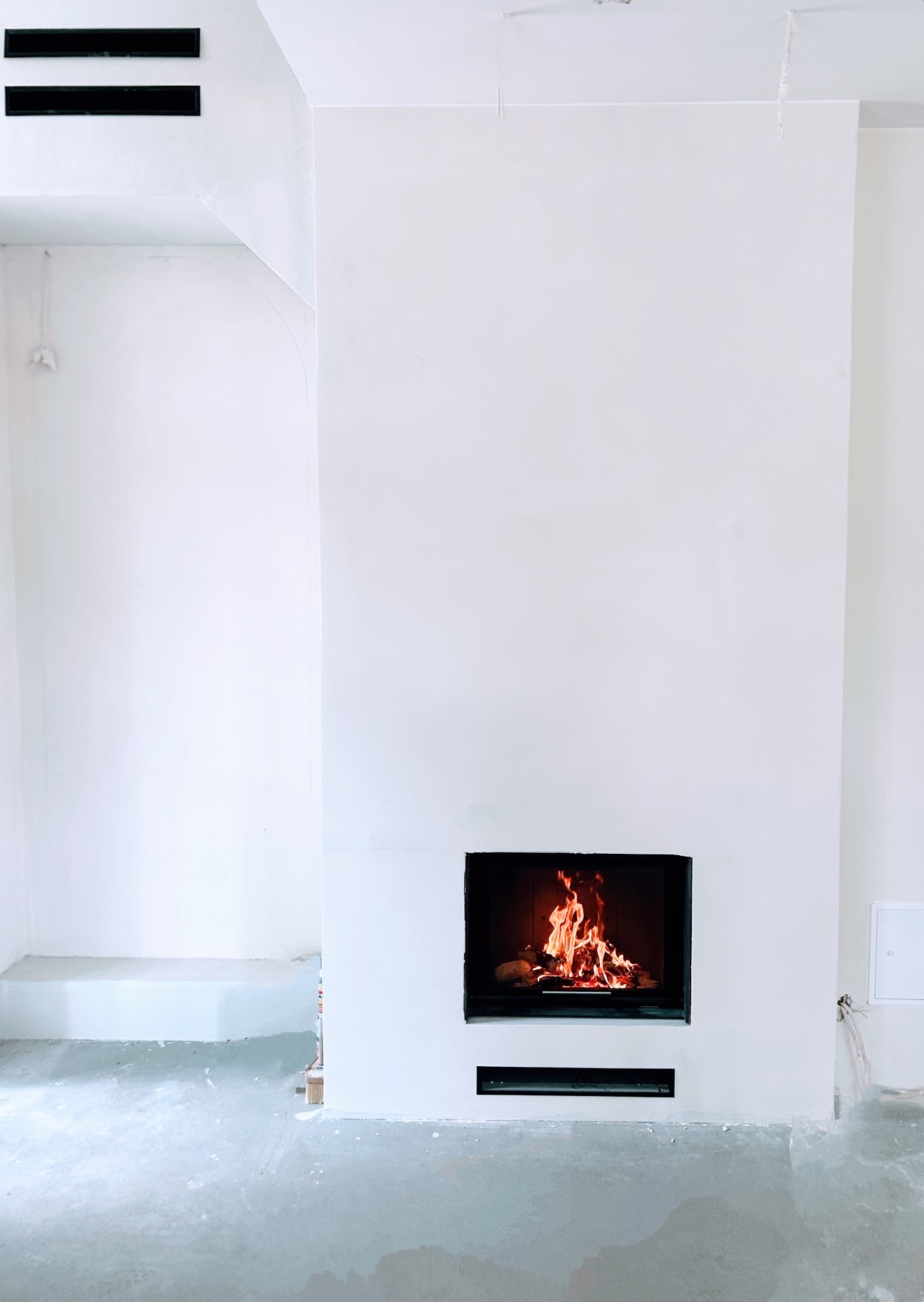 Nowoczesny, minimalistyczny kominek z czarną obudową i paleniskiem w jasnym, surowym wnętrzu. Widoczne detale wykończeniowe i kratki wentylacyjne.