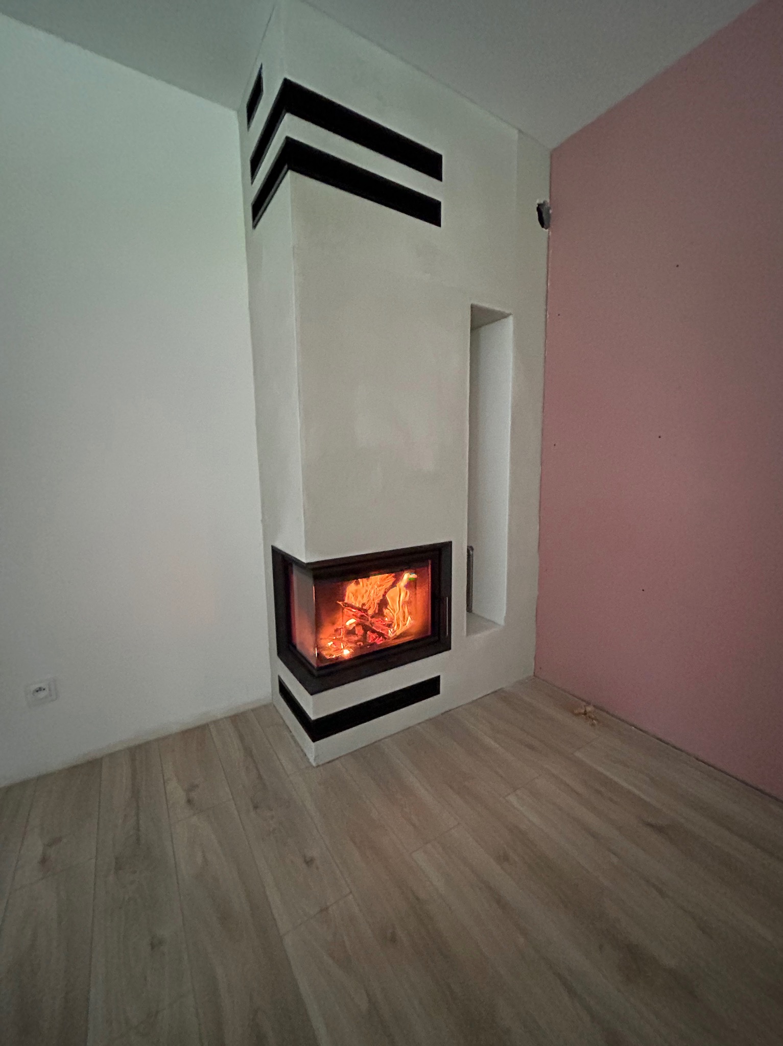 Narożny kominek z paleniskiem, obudowany jasnym tynkiem z czarnymi pasami, w rogu pokoju z panelami podłogowymi i ścianami w kolorze białym i różowym.