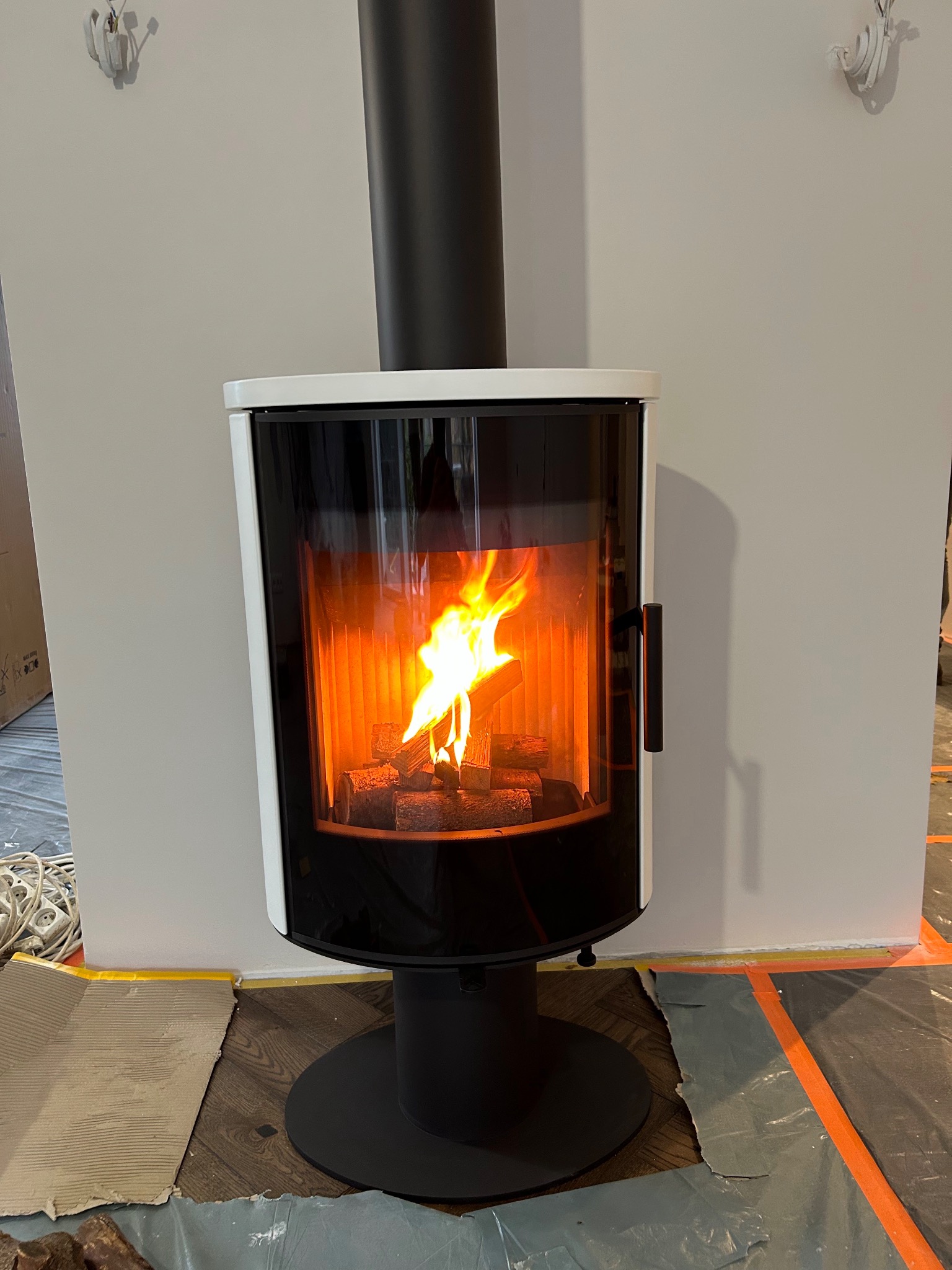 Nowoczesny kominek wolnostojący z paleniskiem, otoczony białym obramowaniem i czarnym wykończeniem, z widocznym płomieniem i przygotowanym zabezpieczeniem podłogi.