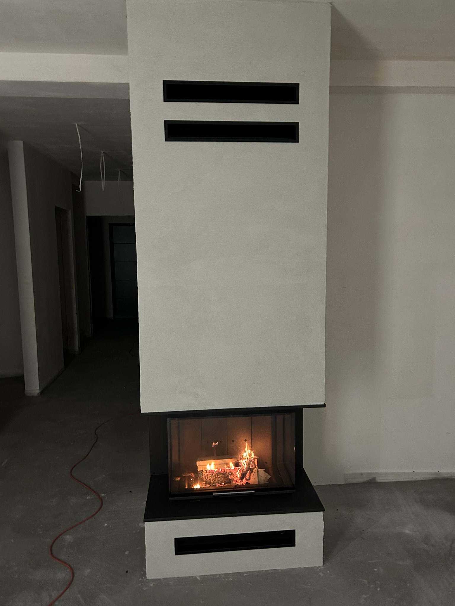 Nowoczesny kominek z paleniskiem w surowym wnętrzu. Minimalistyczny design z czarnymi akcentami i płomieniami. Widoczne niedokończone wykończenie podłogi.