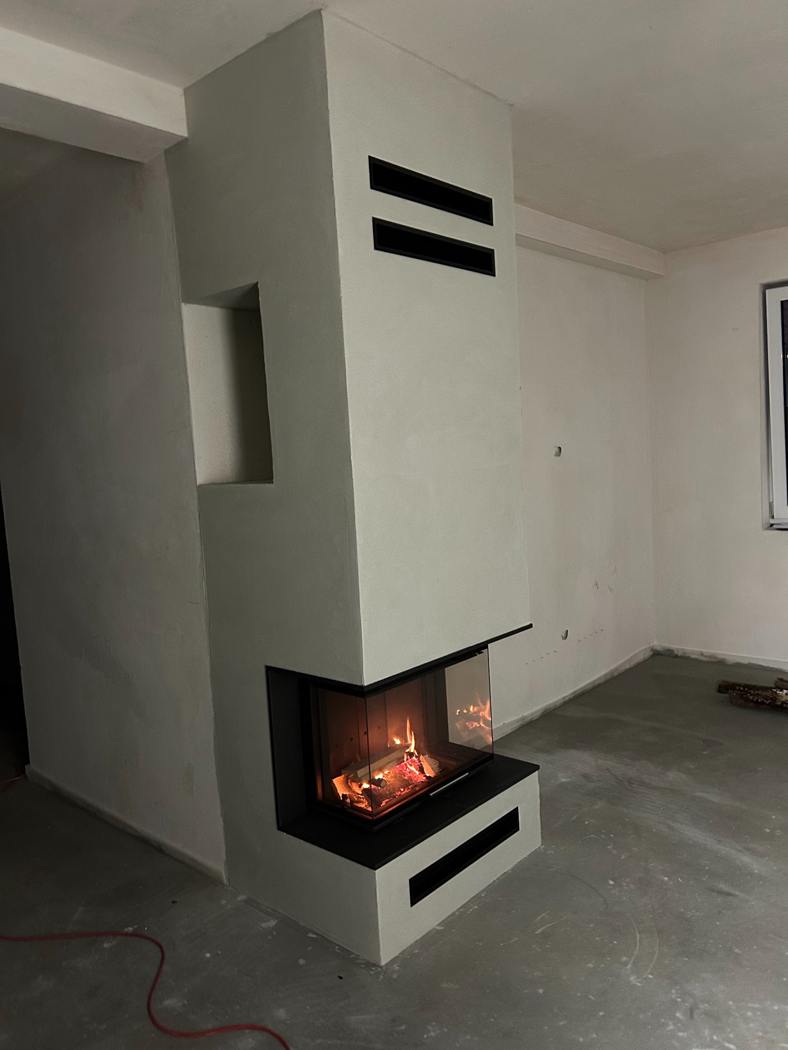 Nowoczesny kominek narożny z paleniskiem, obudowany jasnym tynkiem strukturalnym i czarnymi akcentami. Widoczne płomienie i surowy beton na podłodze.