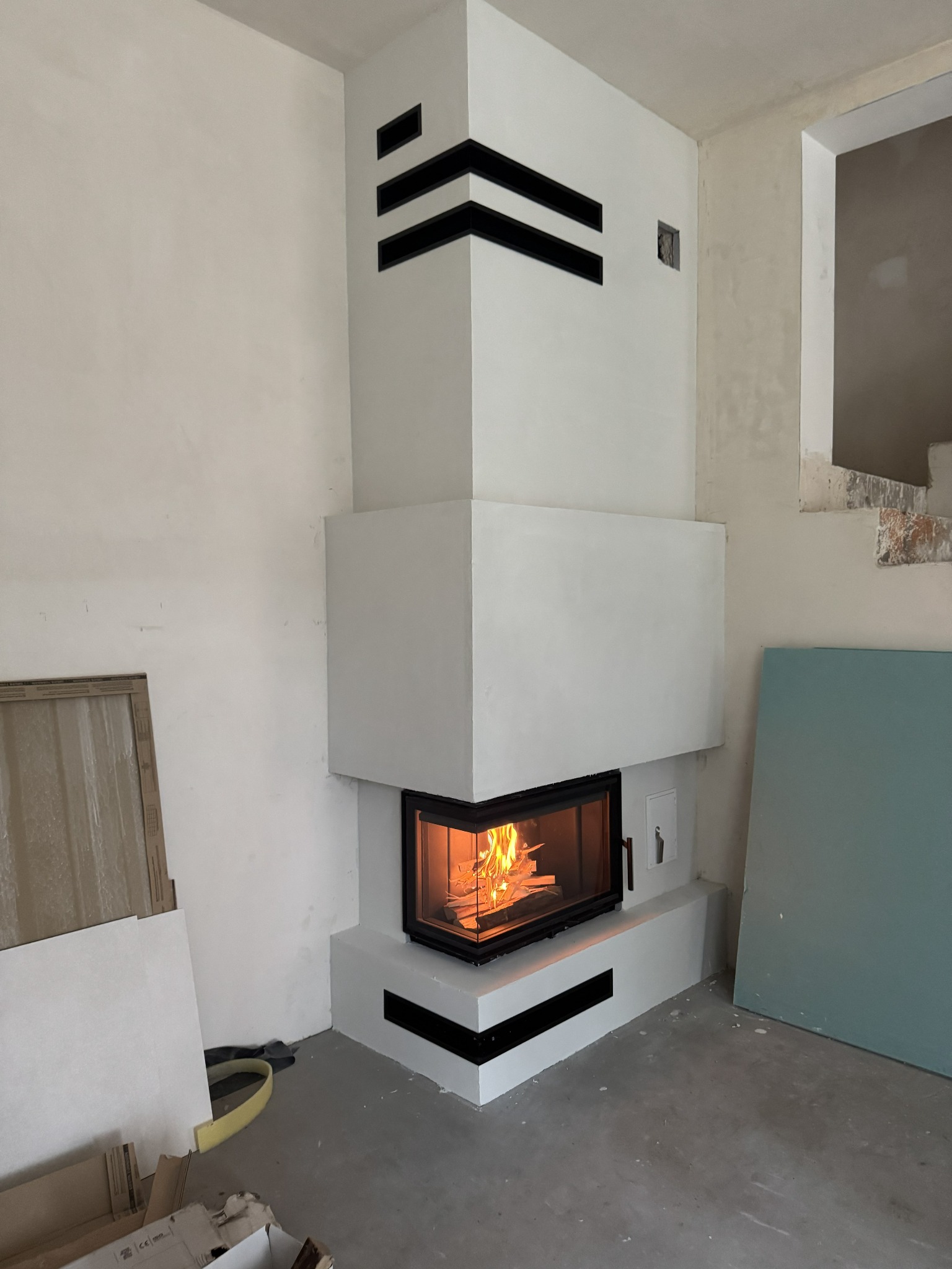 Nowoczesny kominek narożny z czarnymi akcentami, płomienie widoczne przez szybę, surowe wnętrze w trakcie wykończenia, płyty gipsowe w tle.