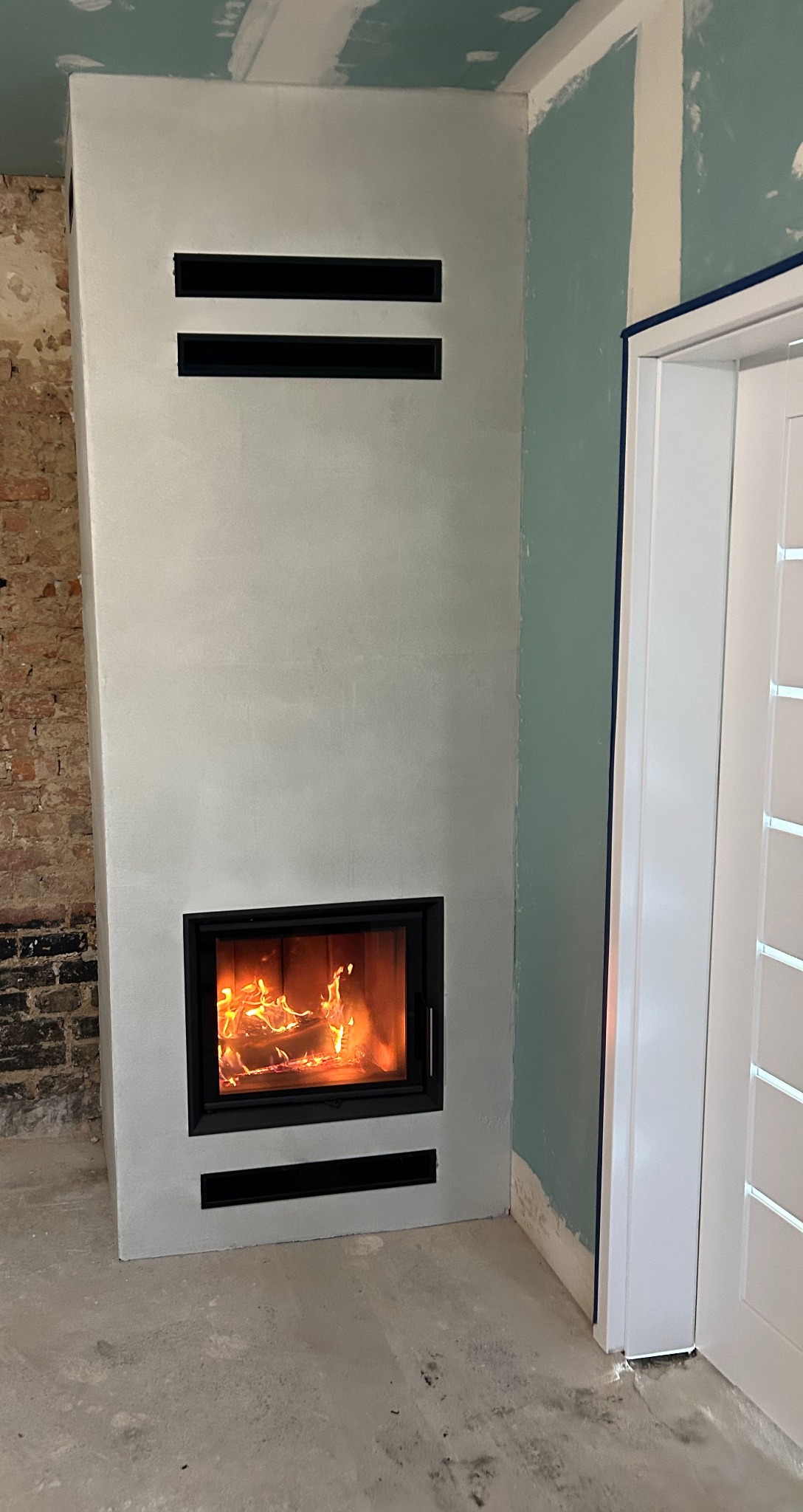 Nowoczesny kominek z paleniskiem w czarnej ramie, w surowym wnętrzu z fragmentem ceglanej ściany i niepomalowanymi ścianami. Widoczne kratki wentylacyjne.