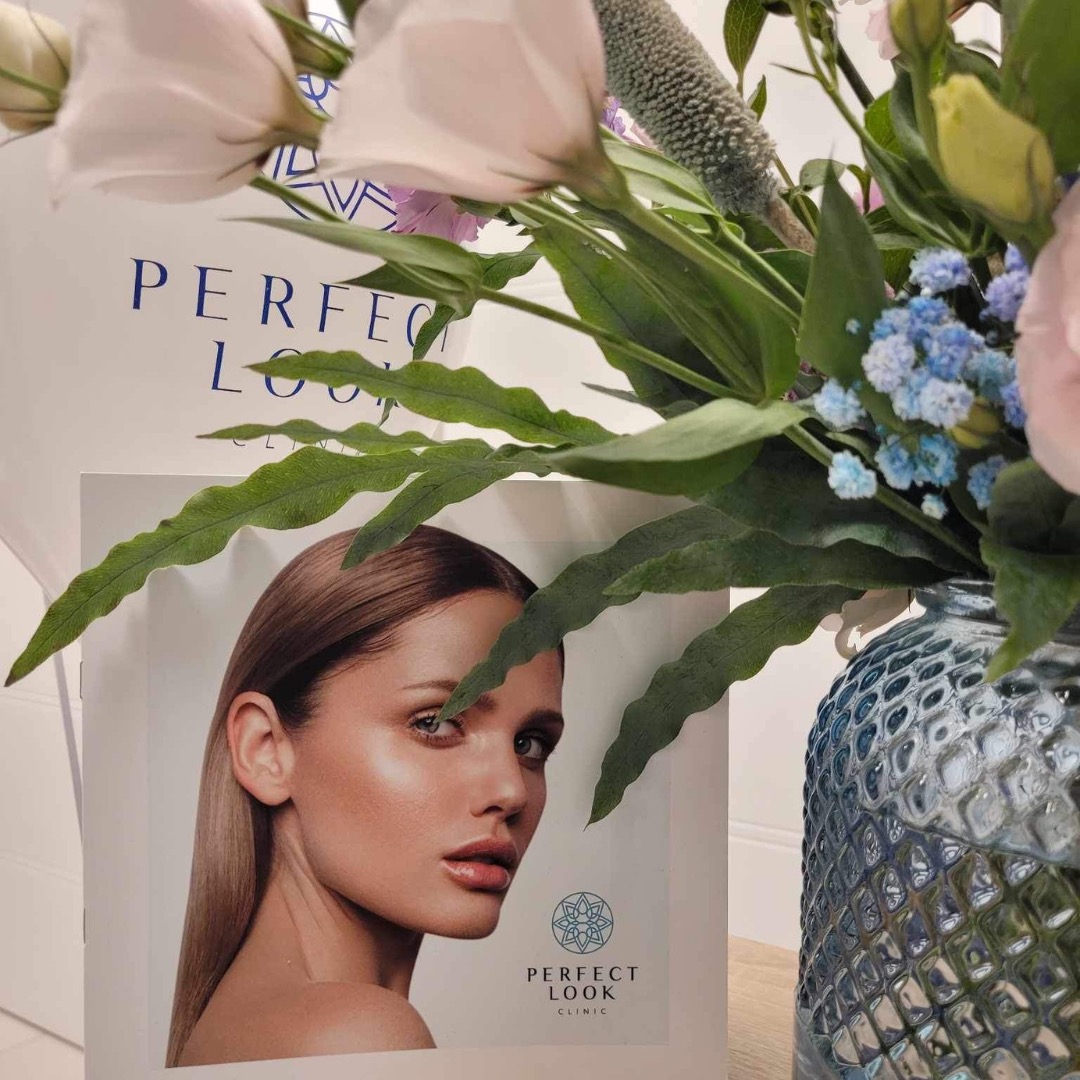 Ujęcie z bliska: ulotka Perfect Look Clinic z portretem kobiety o gładkiej cerze, ustawiona obok niebieskiego wazonu z kwiatami, tworząca estetyczną kompozycję.