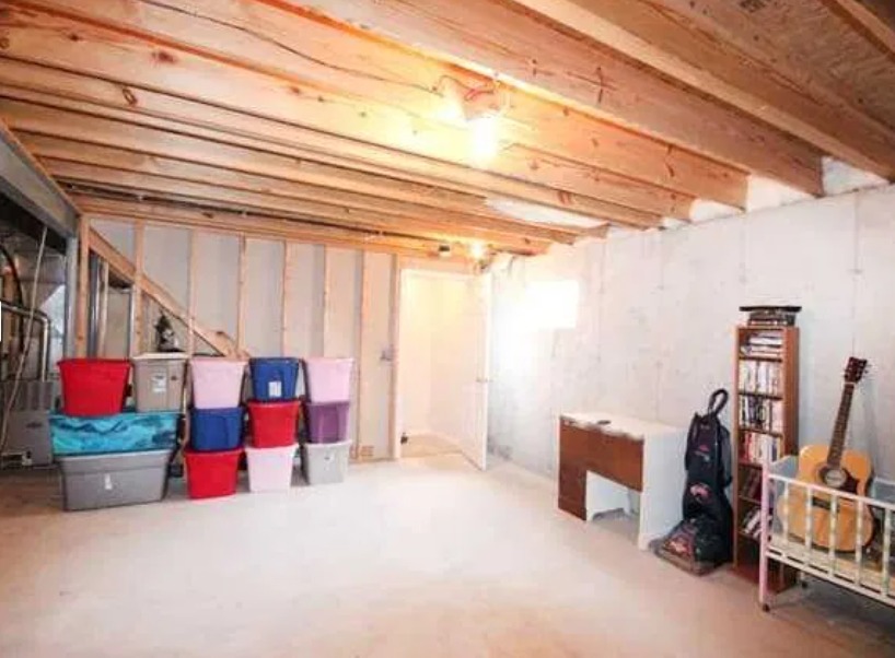 Piwnica przed opróżnianiem: widoczne liczne plastikowe pojemniki, regał z książkami, gitara oparta o łóżeczko dziecięce, odkurzacz workowy, surowy drewniany strop i częściowo wykończone ściany.