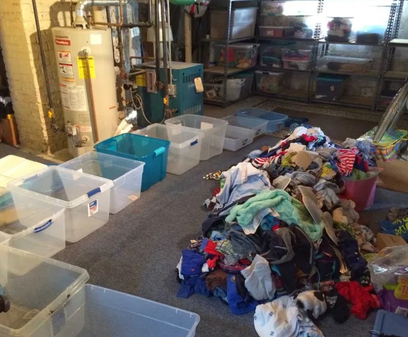 Piwnica z stertą ubrań na dywanie i rzędem plastikowych pojemników do przechowywania, bojler i piec w tle.