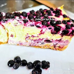 Zakręcona Babeczka - Kawałek sernika z jagodami na białym talerzu, z widocznym przekrojem warstw ciasta i dekoracją z owoców. Kilka jagód leży obok ciasta.