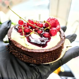 Zakręcona Babeczka - Mini czekoladowa tarta z kremem, wiśniami i maliną, trzymana w dłoni w czarnej rękawiczce.