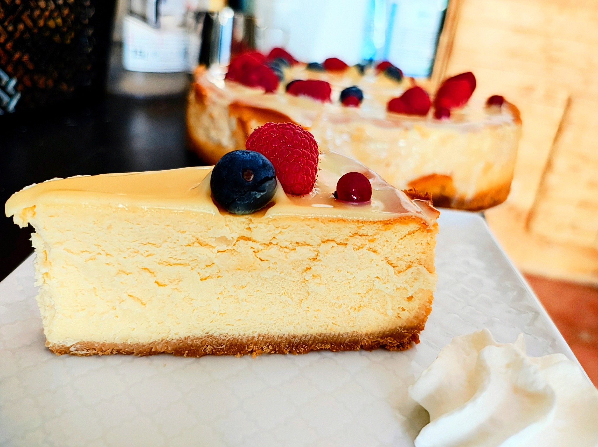 Kremowy sernik nowojorski z owocami: maliną, borówką i czerwoną porzeczką, na białym talerzu z kleksem bitej śmietany, w tle cały sernik.