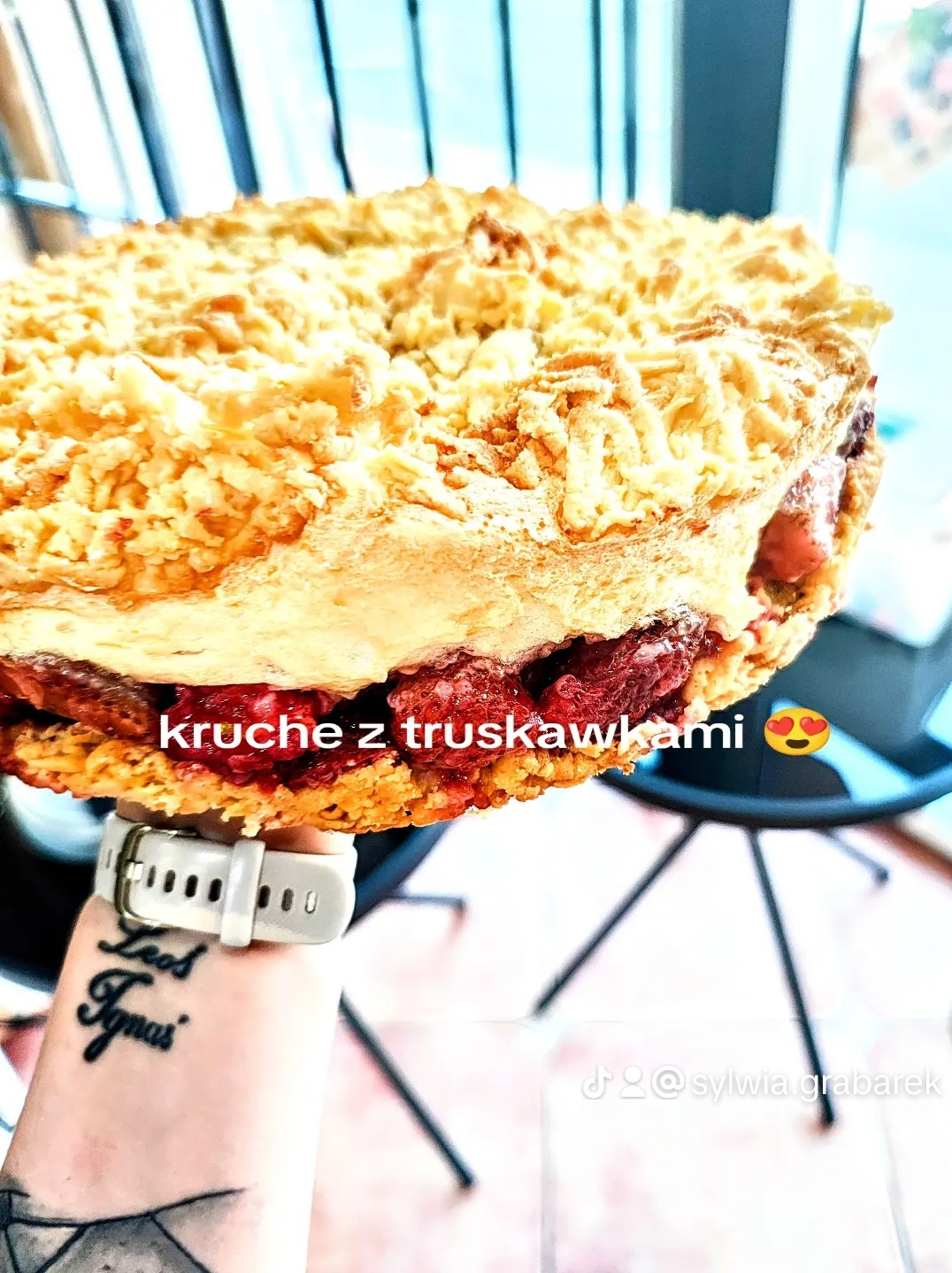 Kruche ciasto z truskawkami, unoszone na dłoni z tatuażem i zegarkiem, widoczne warstwy ciasta i owoców, jasne oświetlenie.