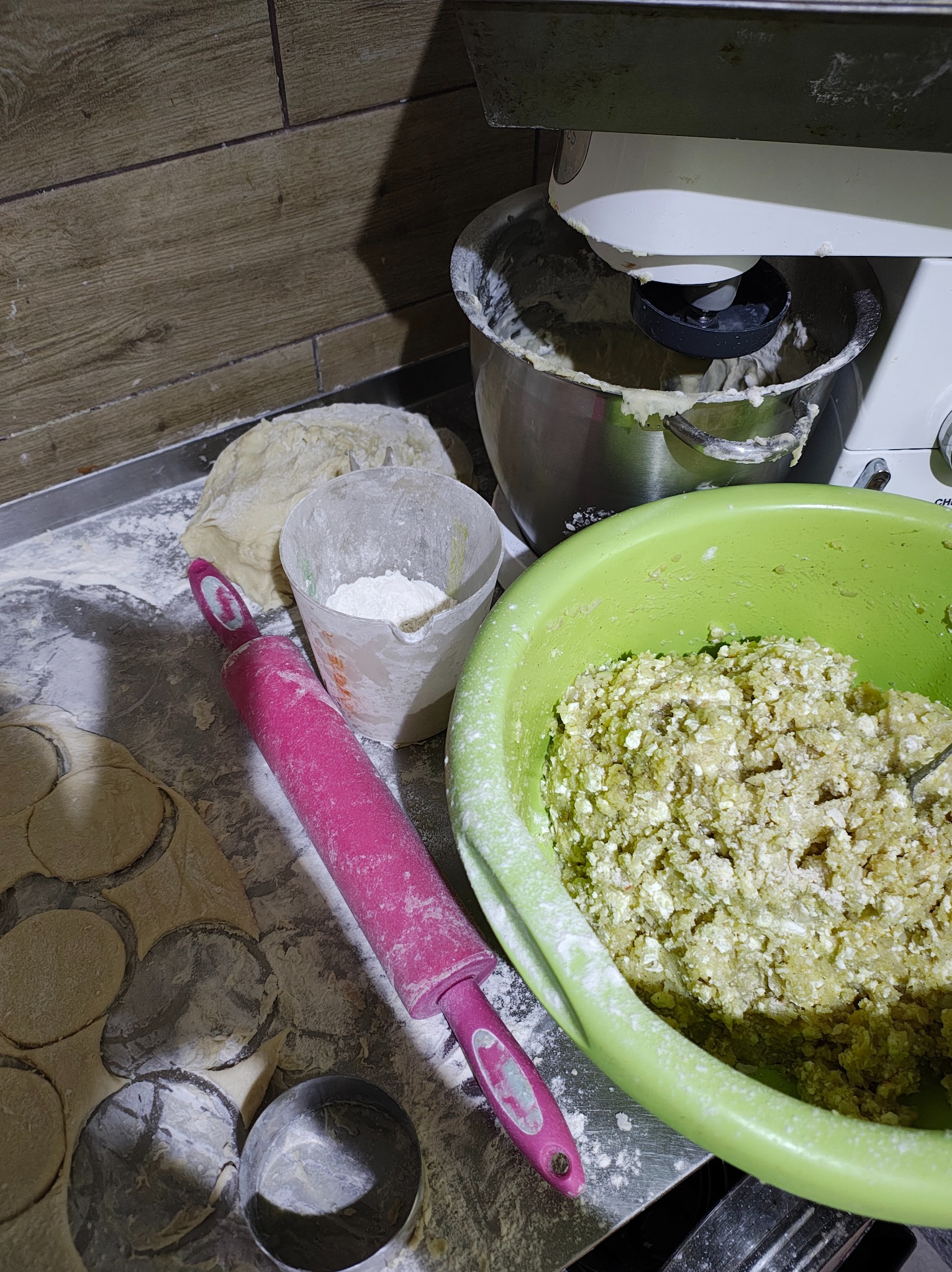 Przygotowywanie ciasta i farszu na pierogi: ciasto w misce miksera, rozwałkowane ciasto z wyciętymi okręgami, różowy wałek, pojemnik z mąką i zielona miska z farszem.