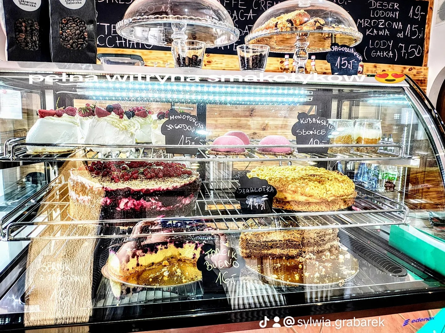 Wnętrze witryny chłodniczej z różnorodnymi ciastami i deserami, w tym bezami z owocami, szarlotką i sernikiem, z cenami wypisanymi na małych tabliczkach, widoczne również menu na tablicy w tle.