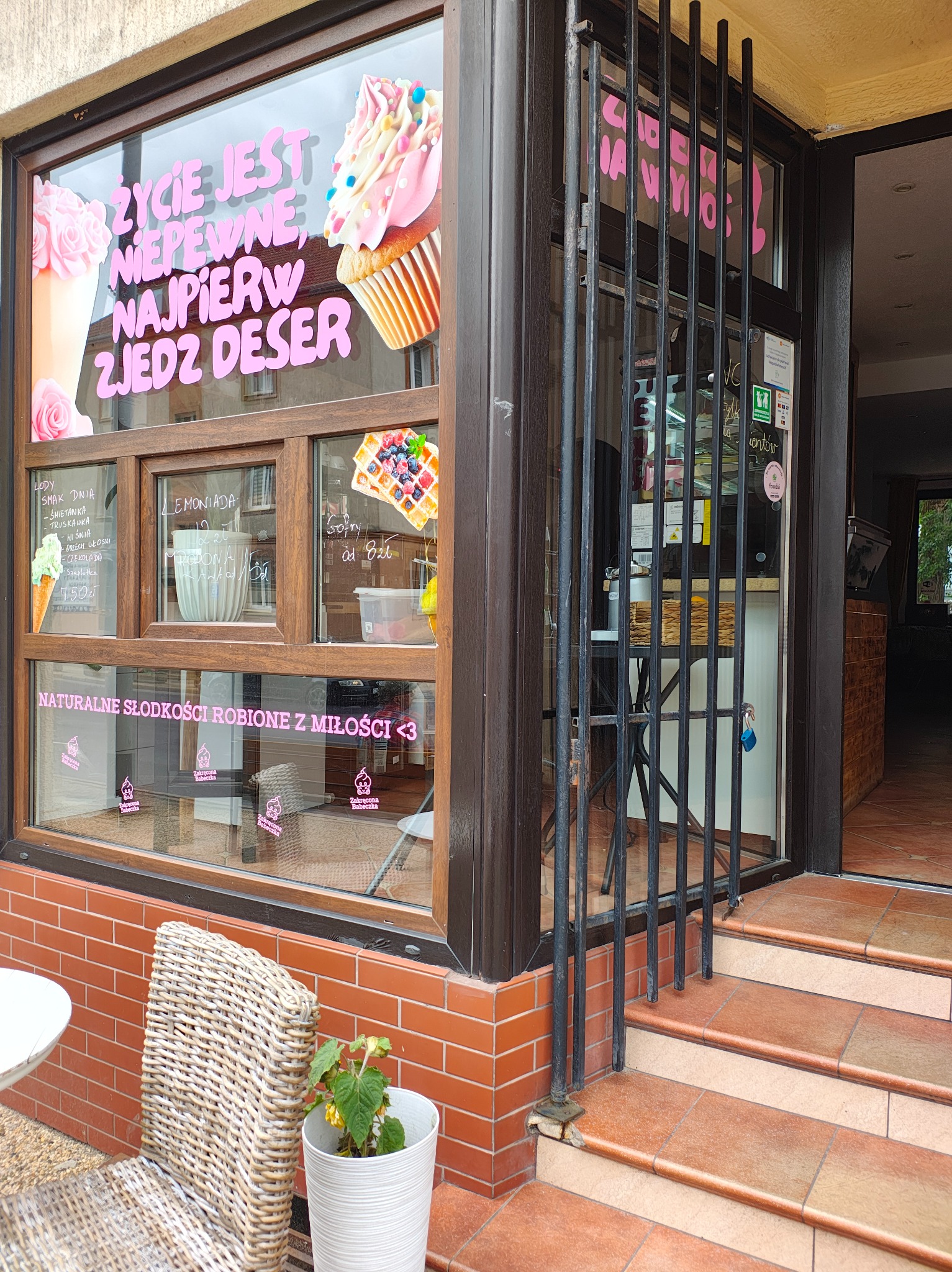 Wejście do kawiarni z oklejonymi szybami, na których widnieją napisy i grafiki reklamujące desery, lody i gofry, a także hasło 'Życie jest niepewne, najpierw zjedz deser', widoczne schody...