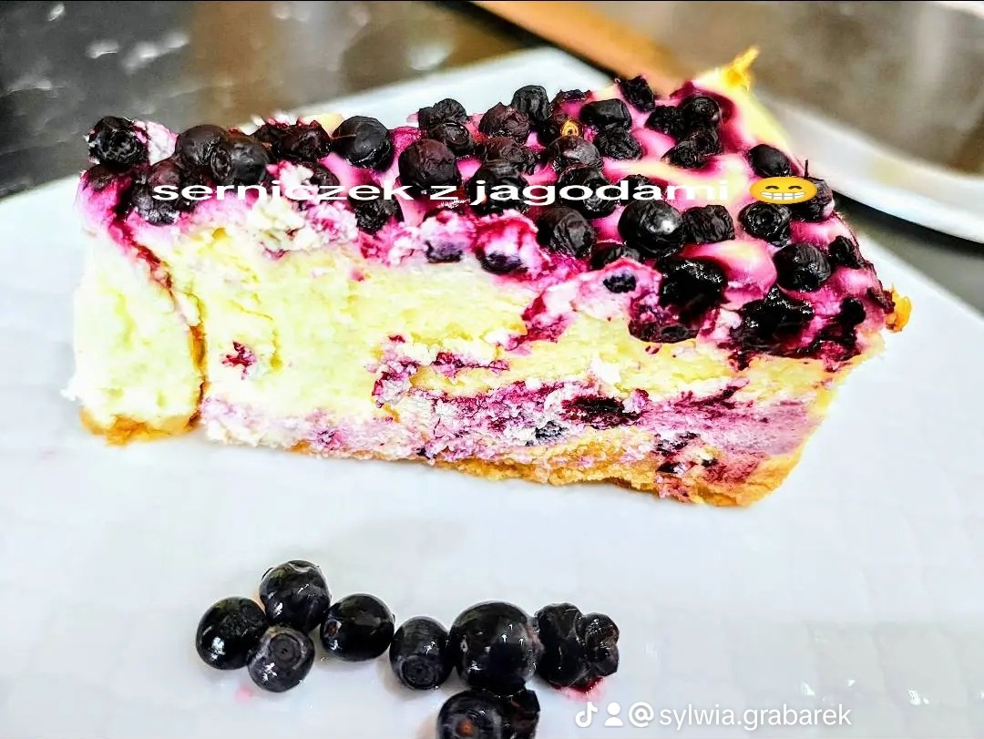 Kawałek sernika z jagodami na białym talerzu, z widocznym przekrojem warstw ciasta i dekoracją z owoców. Kilka jagód leży obok ciasta.