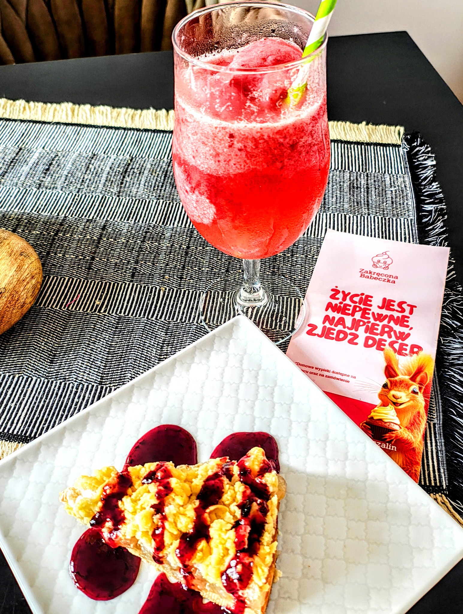 Kawałek ciasta polanego sosem owocowym na białym talerzu obok czerwonego napoju z lodem i słomką oraz ulotki reklamowej Zakręcona Babeczka z wiewiórką.