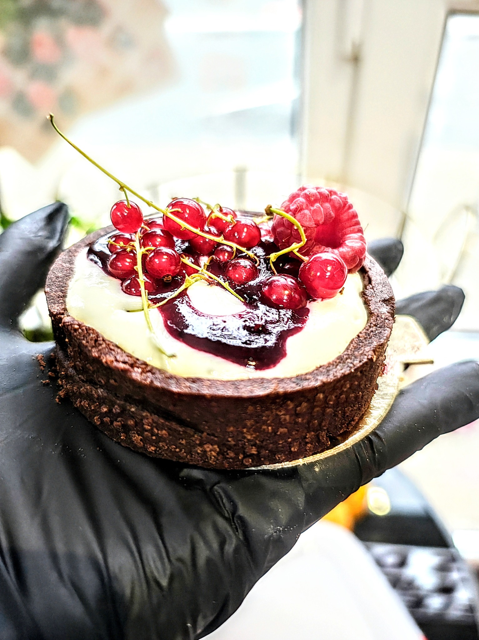 Mini czekoladowa tarta z kremem, wiśniami i maliną, trzymana w dłoni w czarnej rękawiczce.