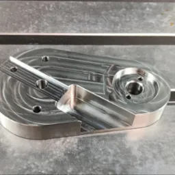 Precyzyjnie wykonany, metalowy element z licznymi otworami, obrobiony w Świdniku, leży na metalowej powierzchni roboczej. Widoczne ślady po obróbce.