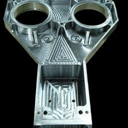 DENMAR MARCIN DENIS - Precyzyjnie obrobiony, srebrny, metalowy element z dwoma okrągłymi otworami i wyfrezowanymi wzorami geometrycznymi na czarnym tle.