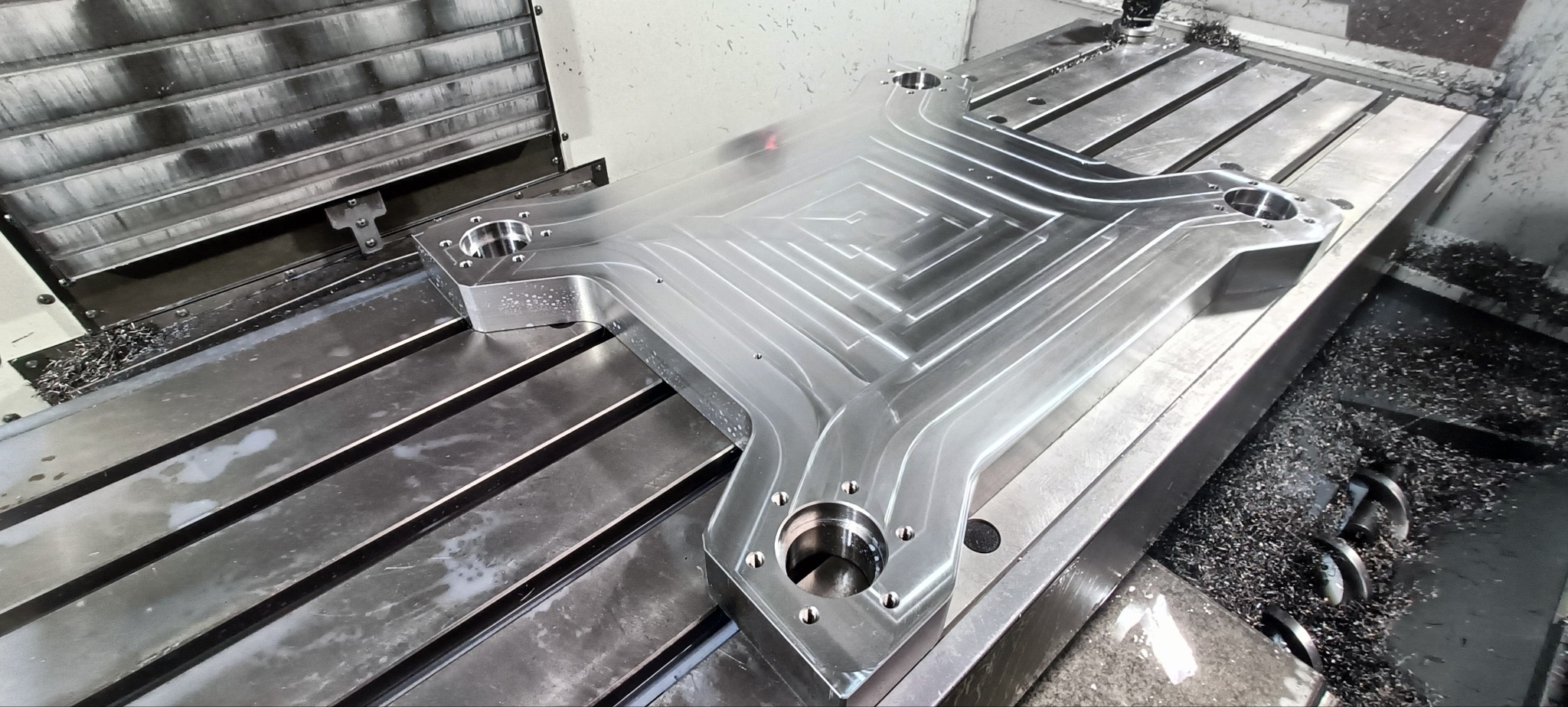 Metalowy element o skomplikowanym kształcie po frezowaniu, leżący na stole frezarki, widoczne wióry i ślady chłodziwa.