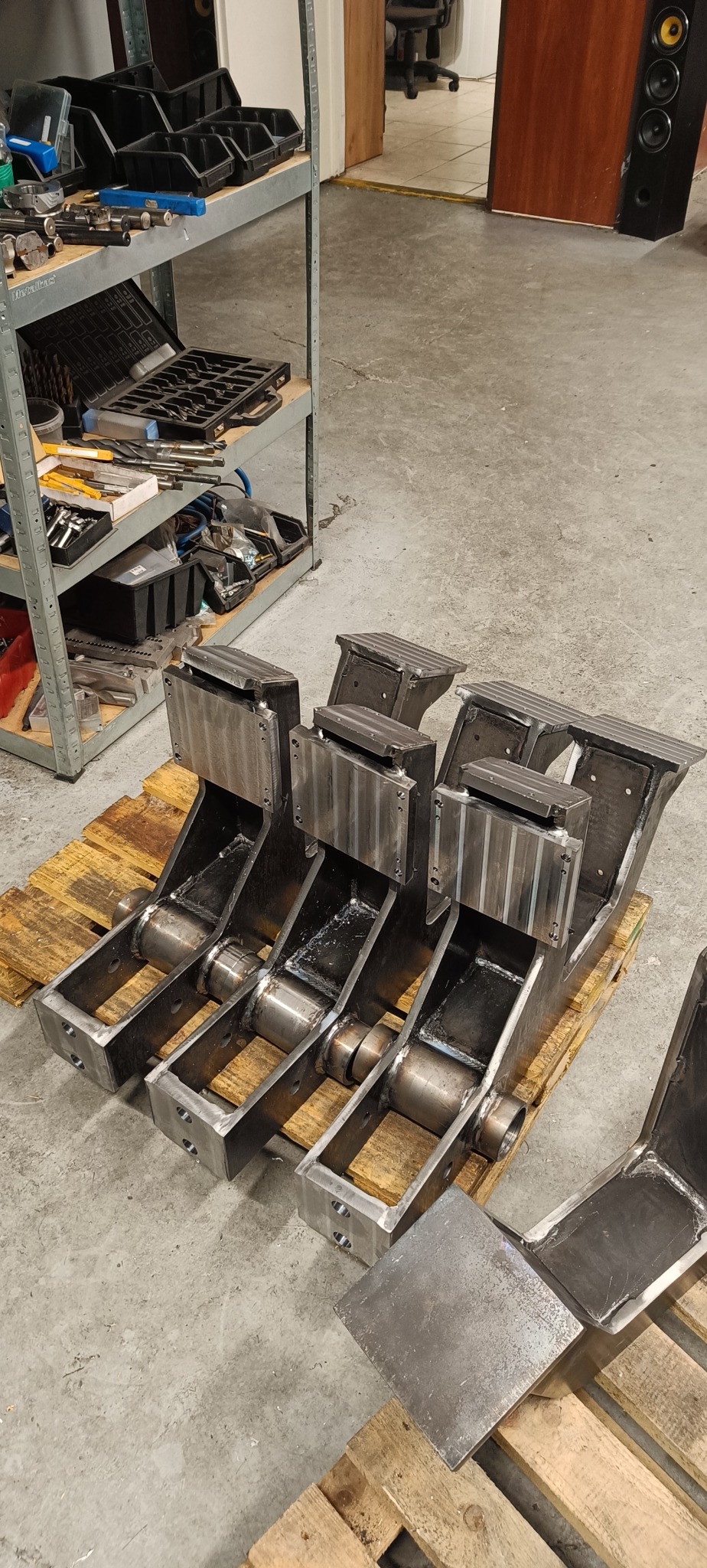 Szereg metalowych, spawanych elementów konstrukcyjnych na palecie w warsztacie, widoczne ślady obróbki i spawania.