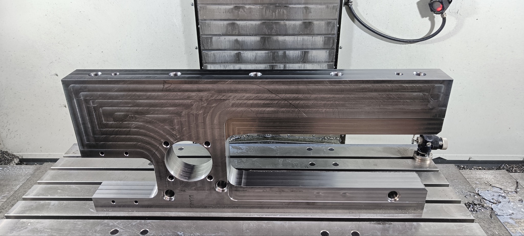 Precyzyjnie obrobiony, metalowy element z licznymi otworami i frezowanymi krawędziami, umieszczony na stole frezarki CNC, widoczne ślady chłodziwa i wiórów.