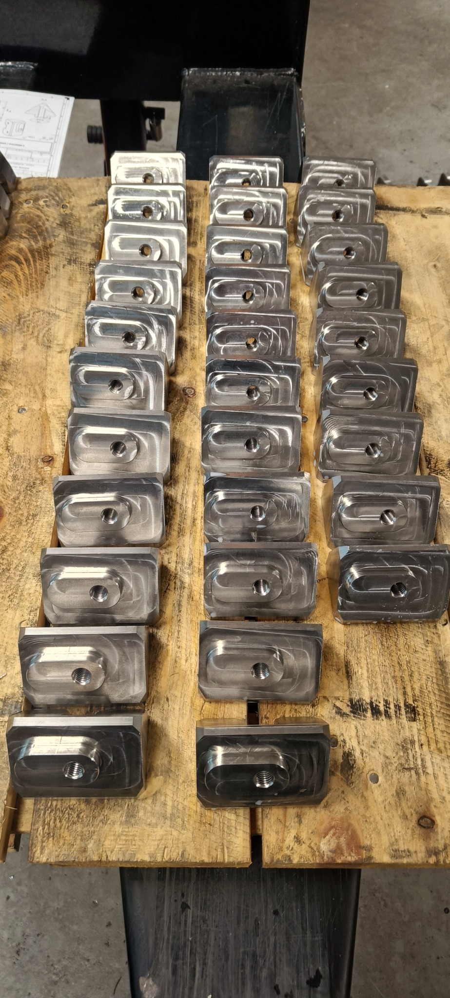 Rząd metalowych, obrobionych elementów z gwintowanymi otworami, ułożonych na drewnianym blacie roboczym z widocznymi śladami użytkowania.